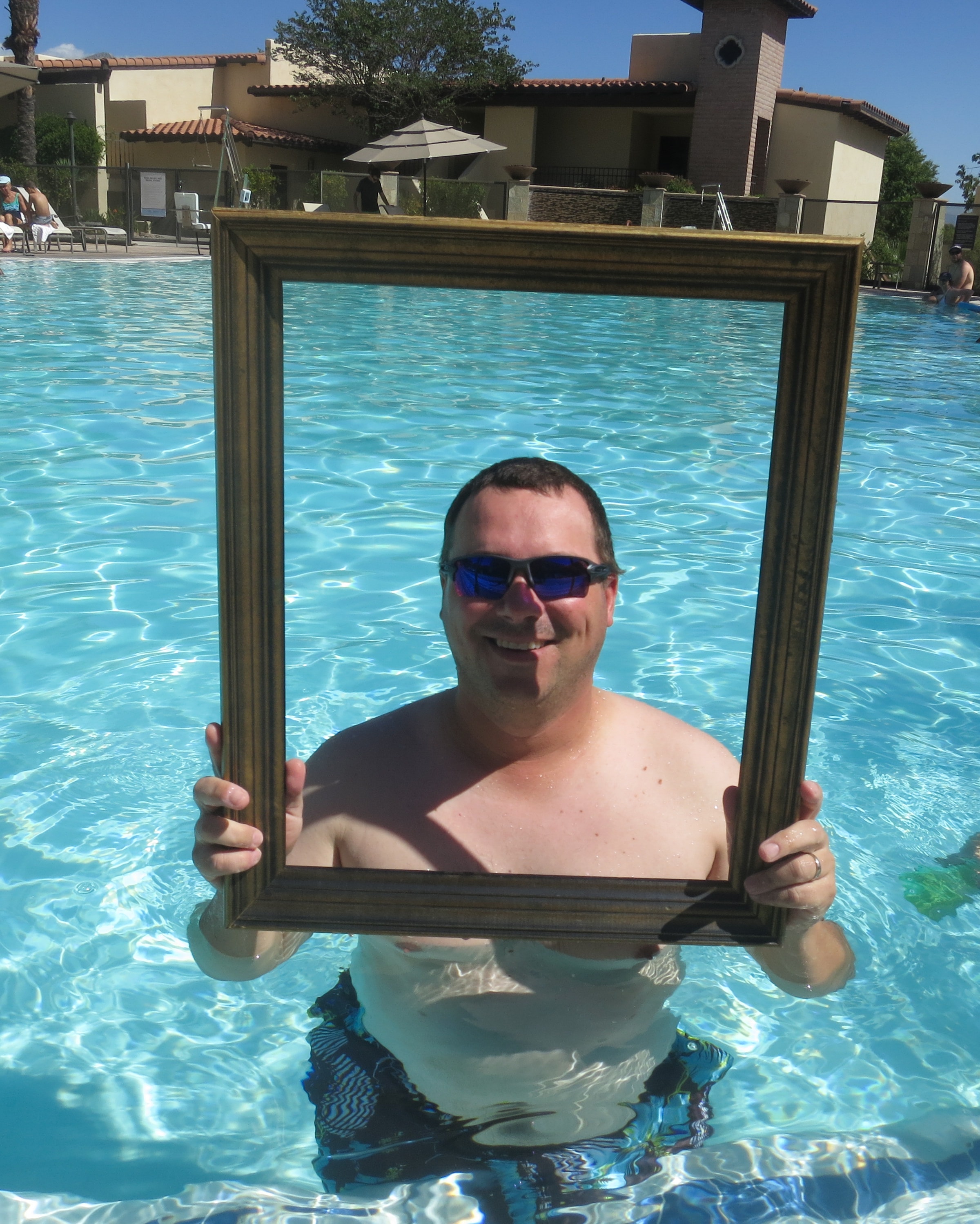 Grinning man in blue mirrored sunglasses holding a gold picture frame while standing chest-deep in a bright turquoise resort swimming pool
