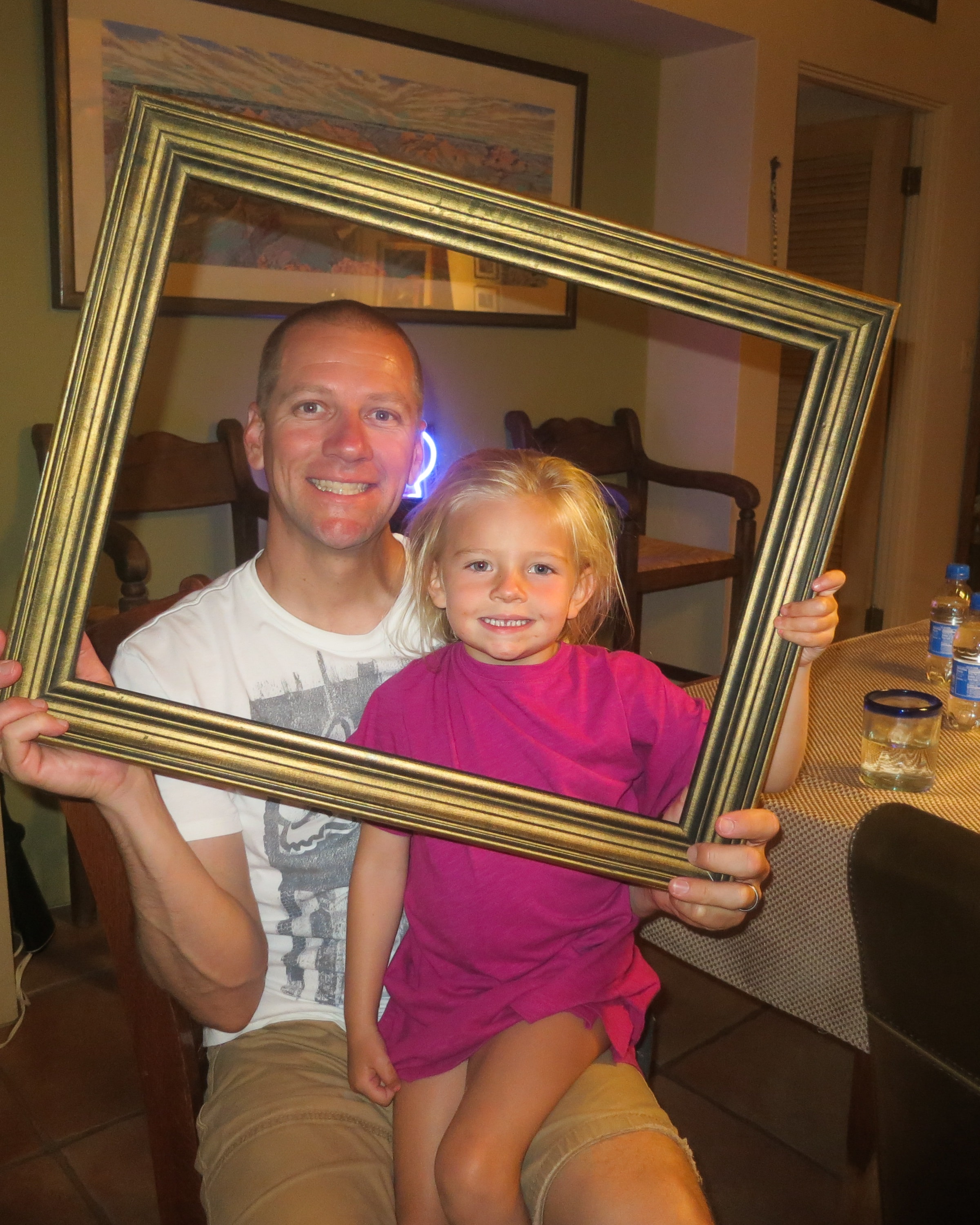 Smiling man in a white graphic t-shirt and a young blonde girl in a pink shirt holding a gold picture frame together at a dining table indoors with Southwest artwork on the walls