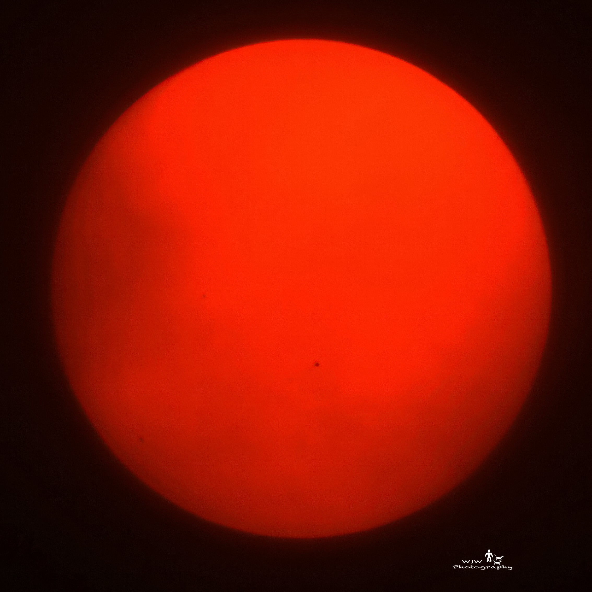 The sun glowing deep red-orange against a black sky with a tiny sunspot visible on its surface