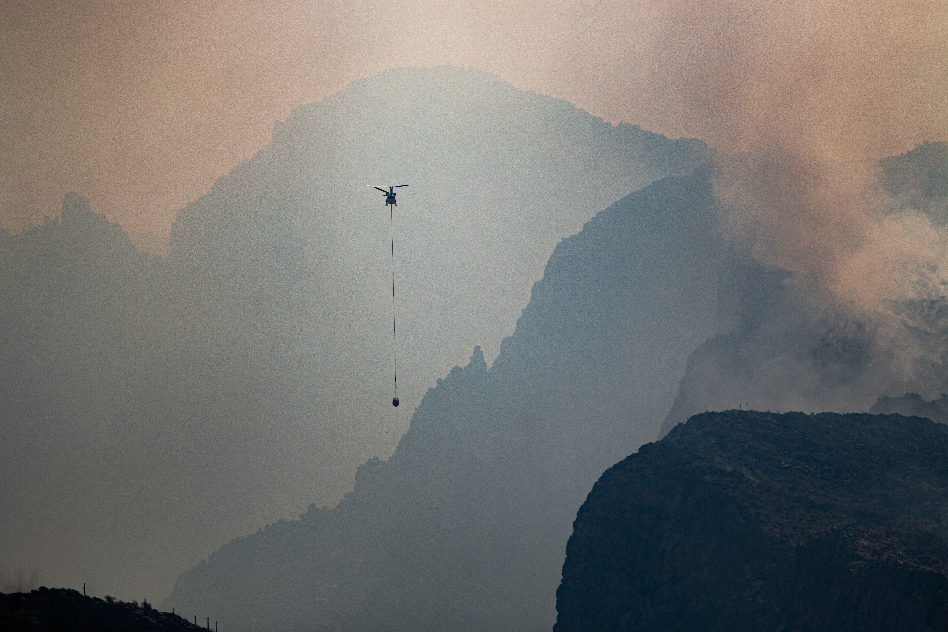 Firefighting helicopter with water bucket suspended over smoke-filled Catalina Mountains during the Bighorn Fire