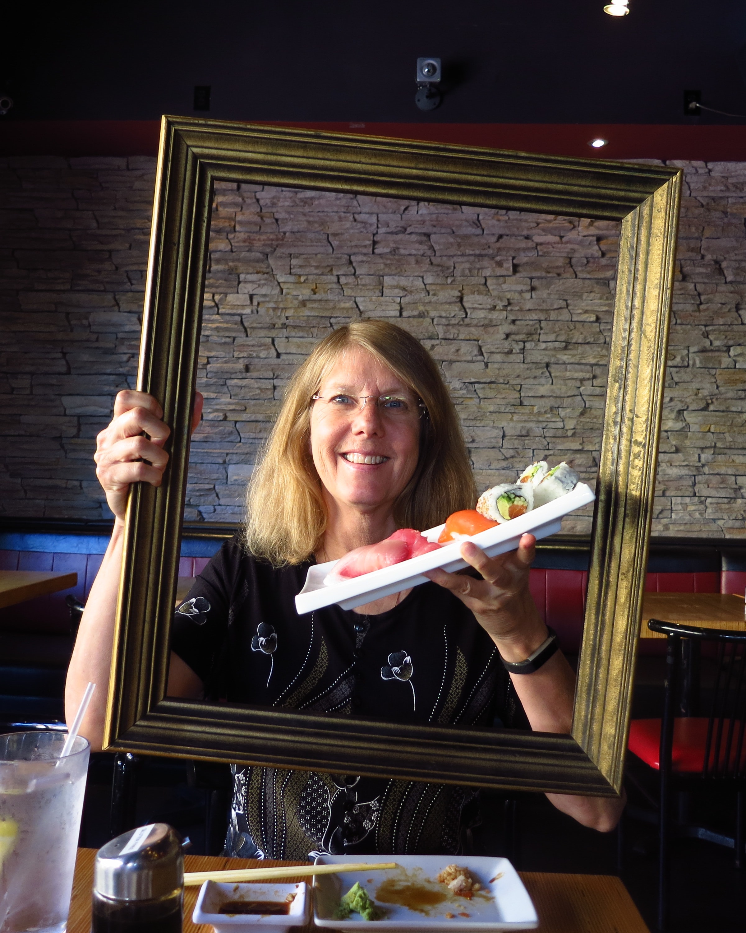 Woman holding a gold picture frame at a sushi restaurant, smiling with a plate of sushi