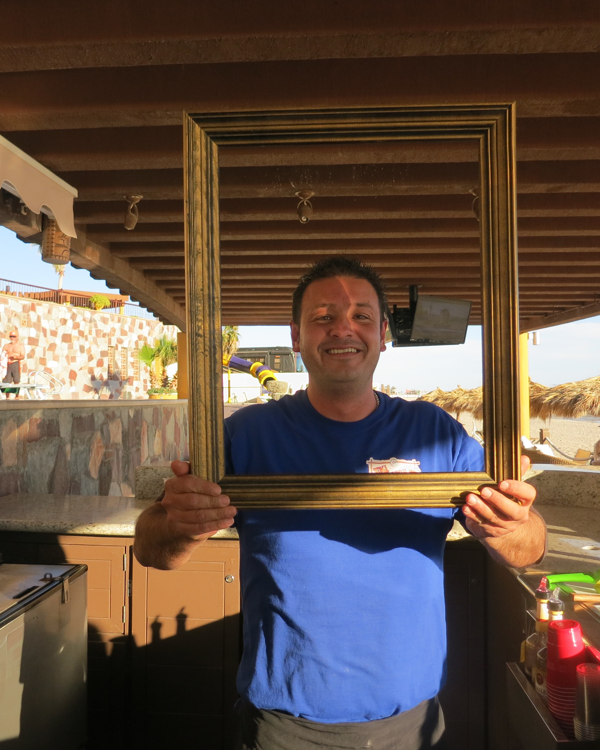Man holding a gold picture frame at a beachside bar, Sea of Cortez in the background