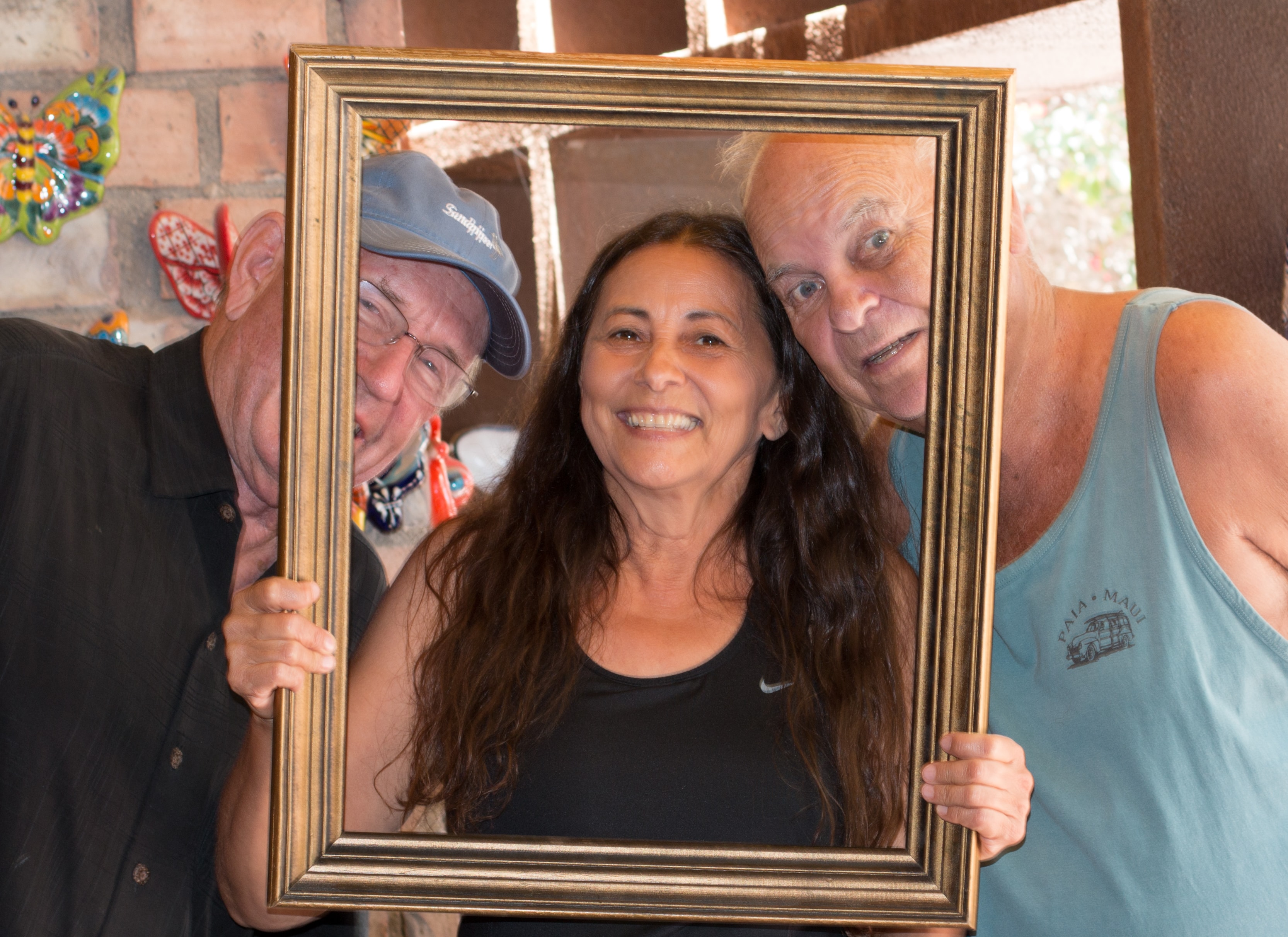 Three people — two men and a woman — crowding together inside a gold picture frame, laughing, with colorful ceramic art on the wall behind them