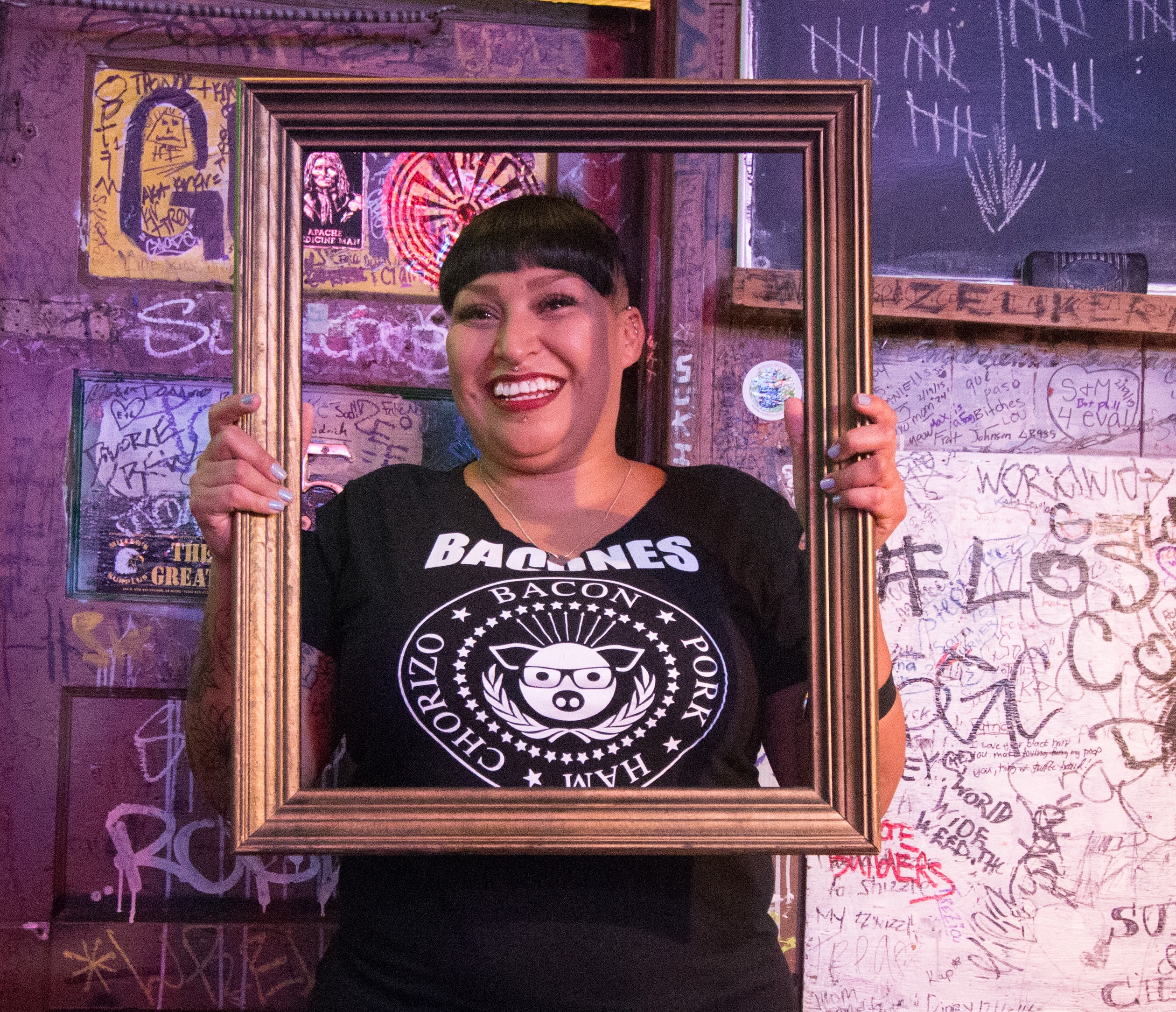 Woman in a Barones t-shirt holding a gold picture frame and smiling wide in front of a graffiti-covered wall