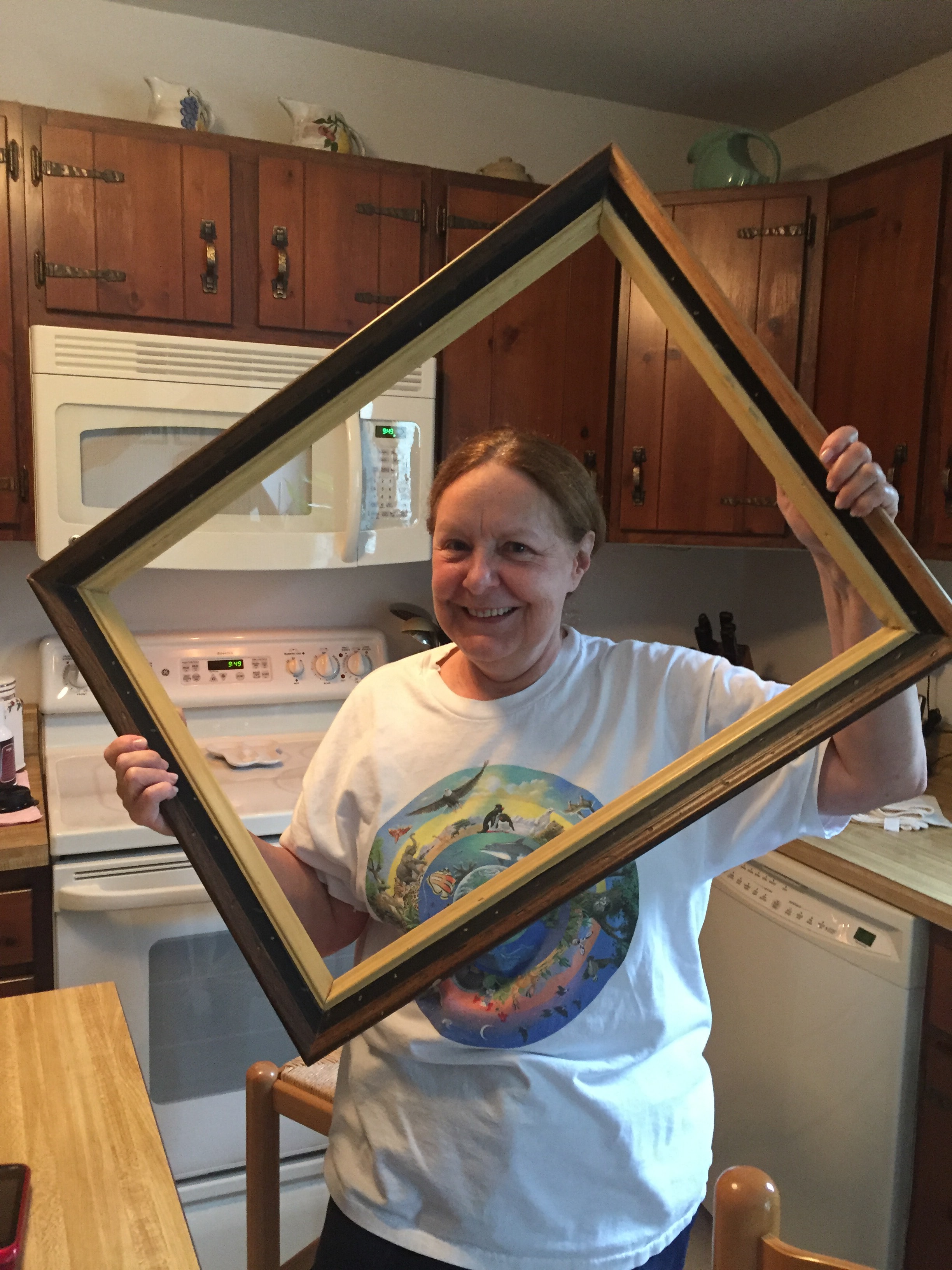 Woman smiling and holding a large wooden picture frame tilted at an angle in a home kitchen