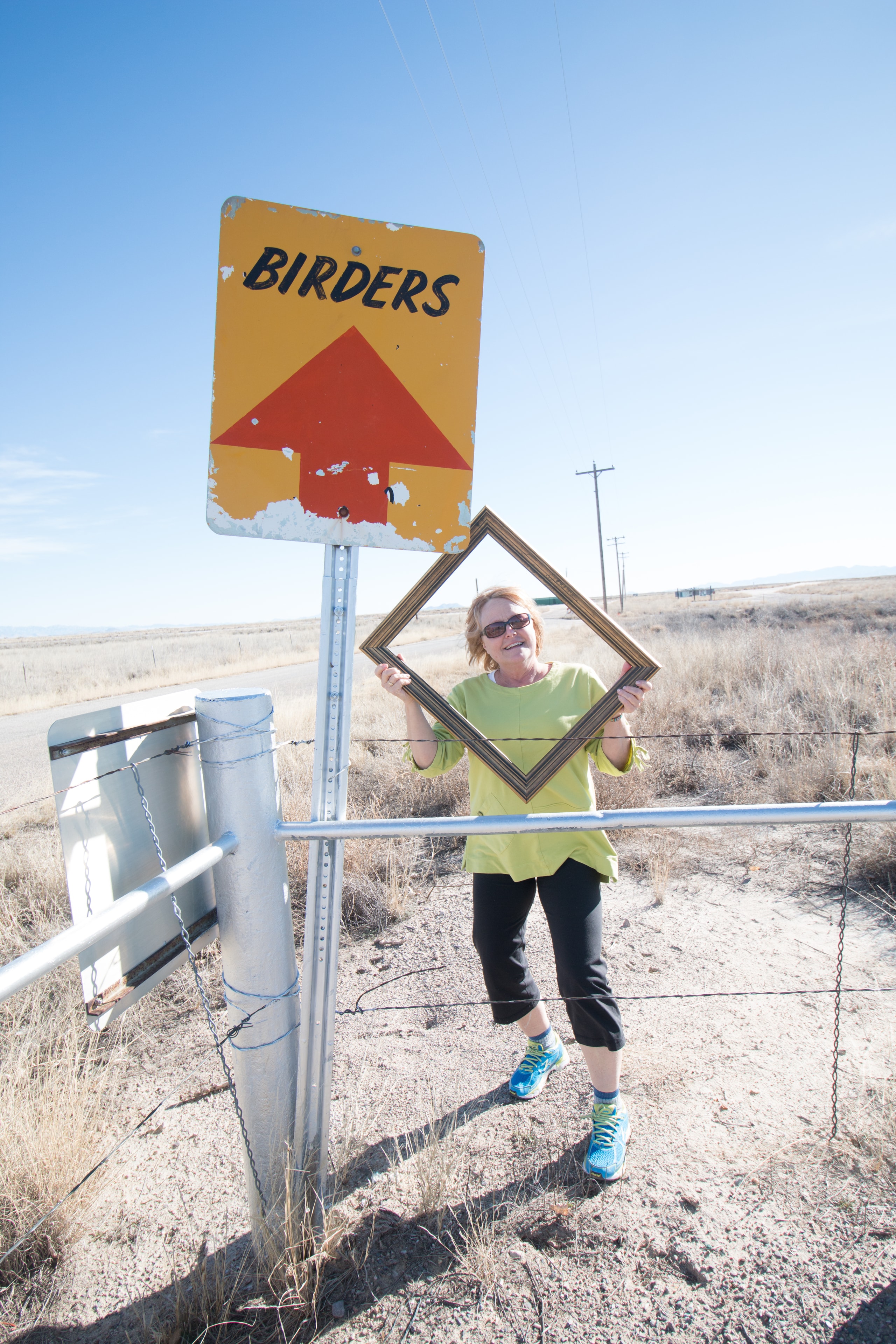 Woman holding a gold picture frame in front of a Birders road sign on an open Arizona grassland