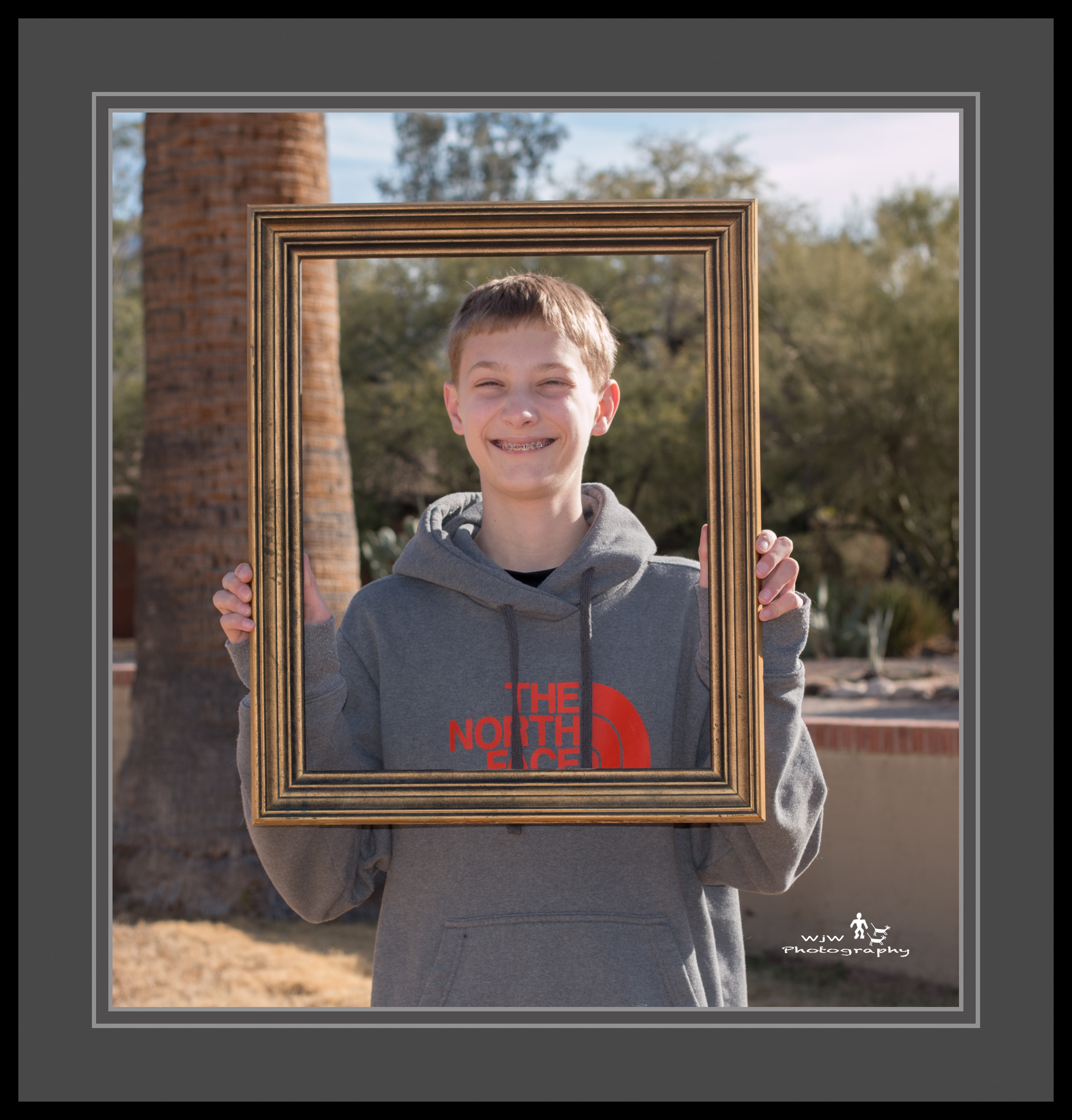 Smiling teenage boy in a North Face hoodie holding a gold picture frame outdoors with palm tree behind him