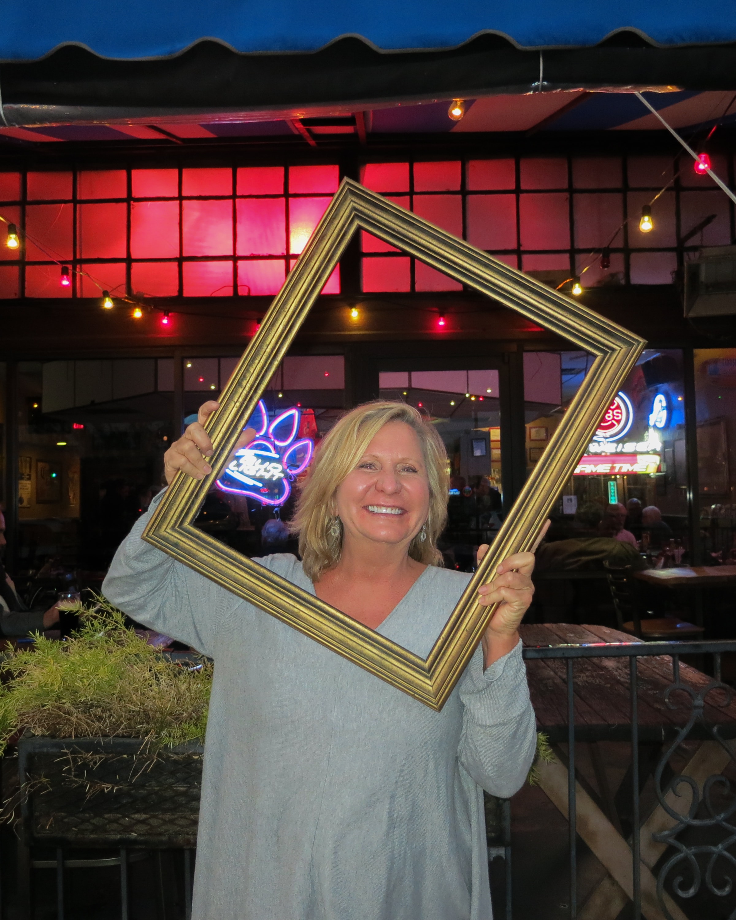 Woman holding a tilted gold picture frame and smiling in front of a neon-lit bar patio at night
