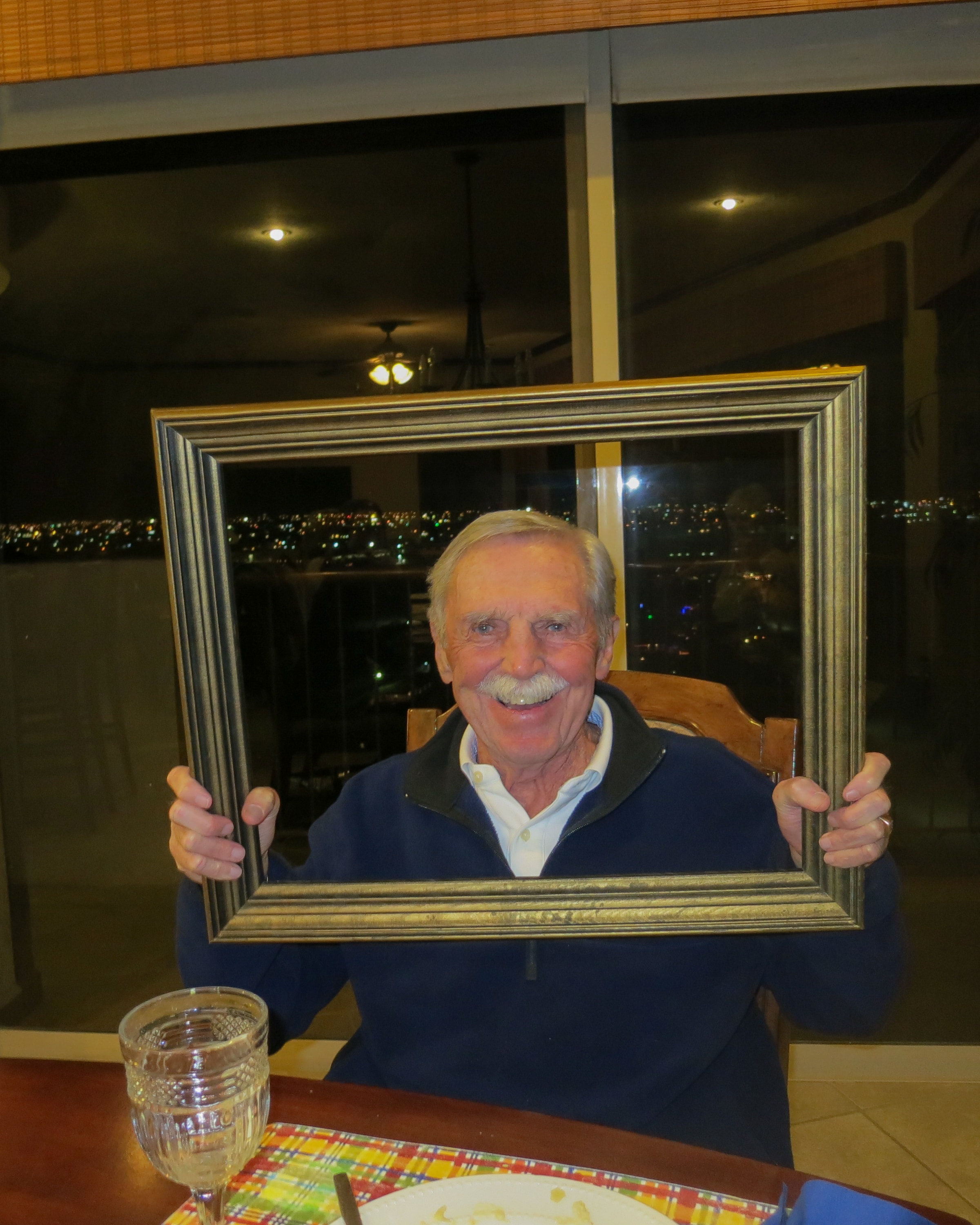 Older gentleman smiling through a silver picture frame at a dinner table with city lights glowing behind him at night