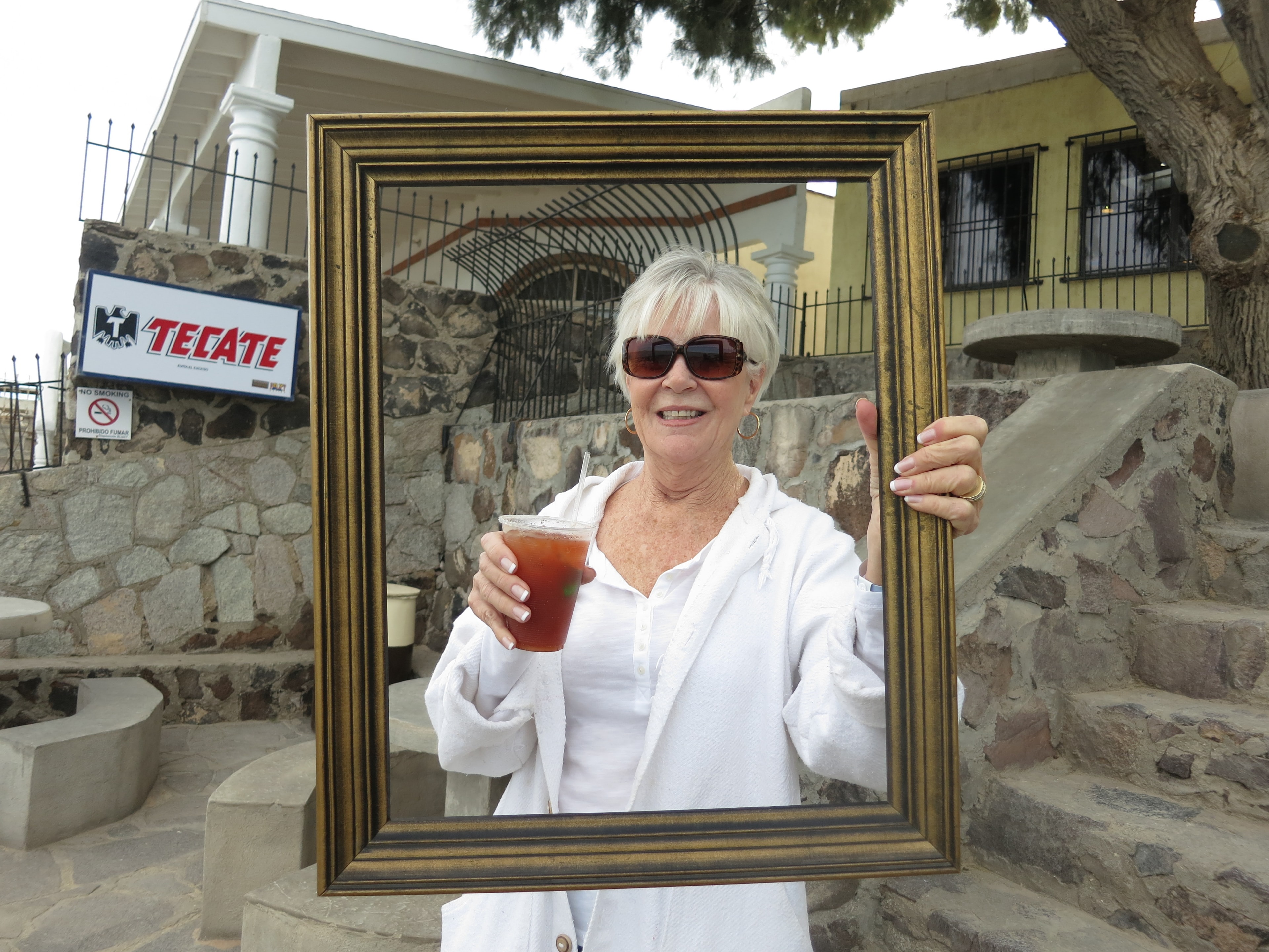Woman in sunglasses holding a gold picture frame and a michelada in front of a Tecate sign at a Mexican cantina