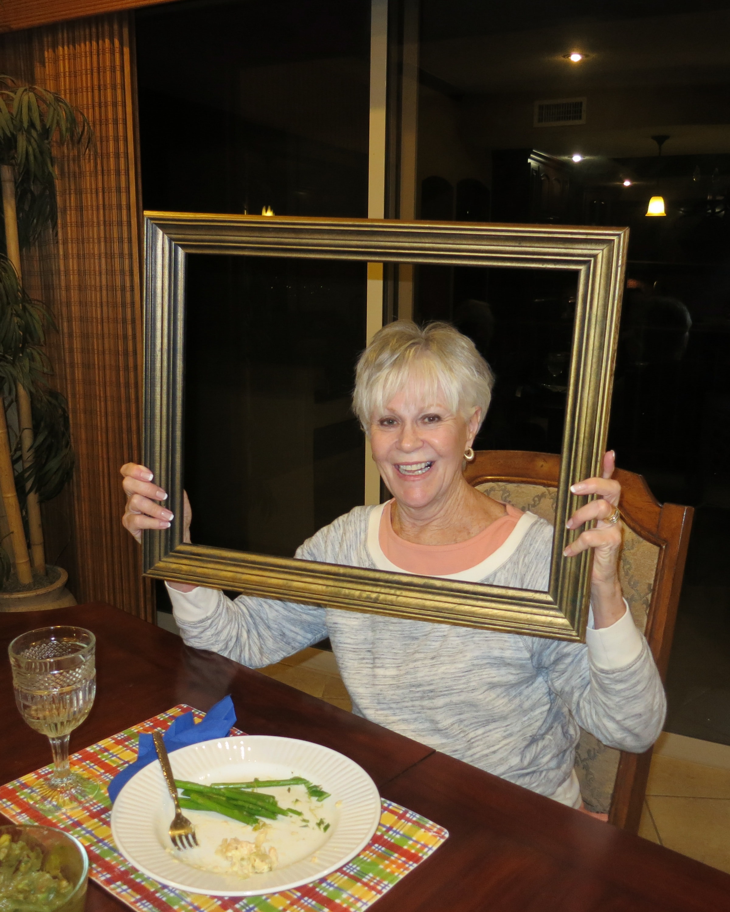 Smiling woman holding a large gold picture frame at a dinner table with a plate of asparagus and a glass of wine
