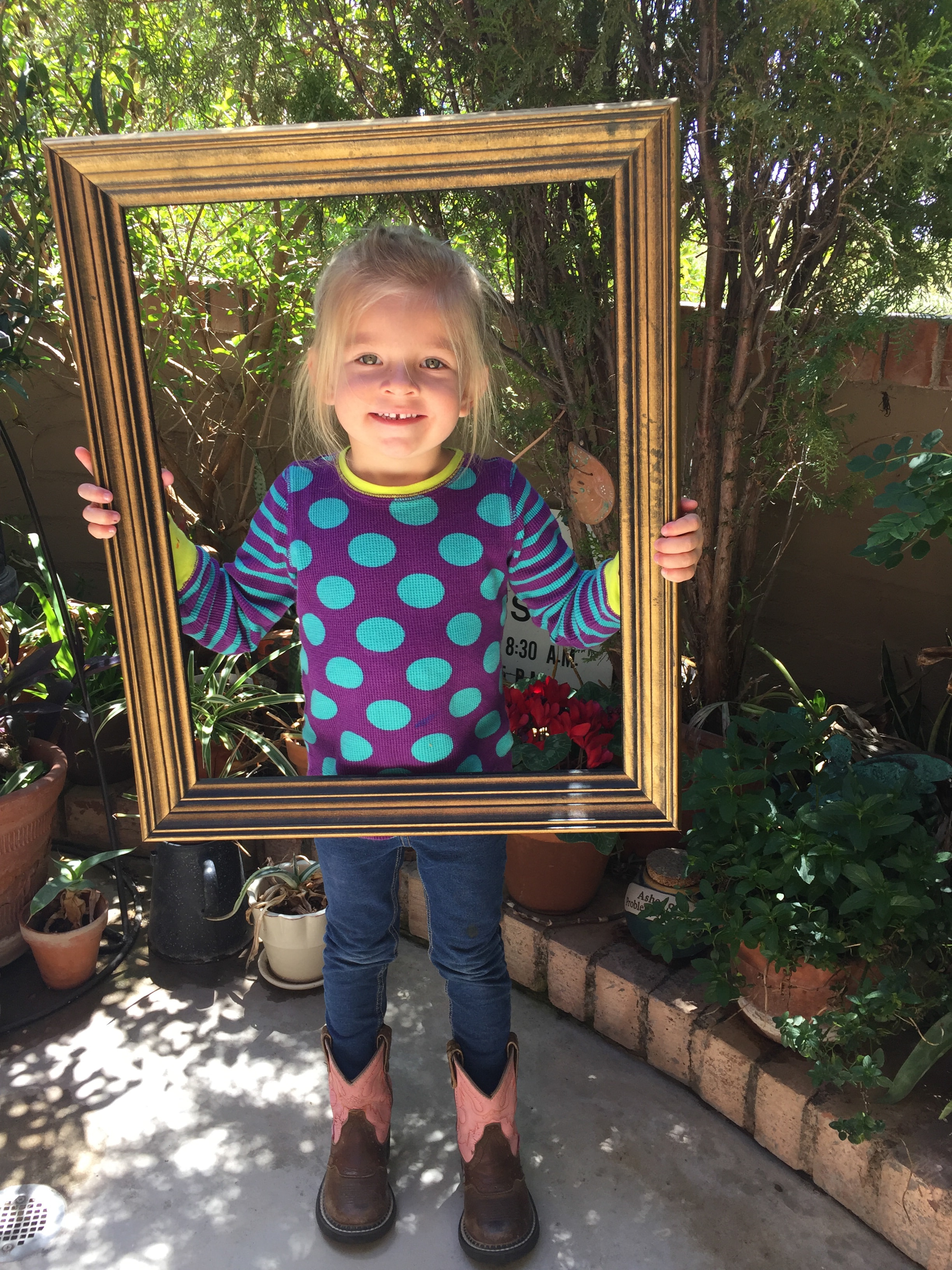 Young blonde girl in a polka-dot shirt and pink cowboy boots holding a large gold picture frame in a garden patio