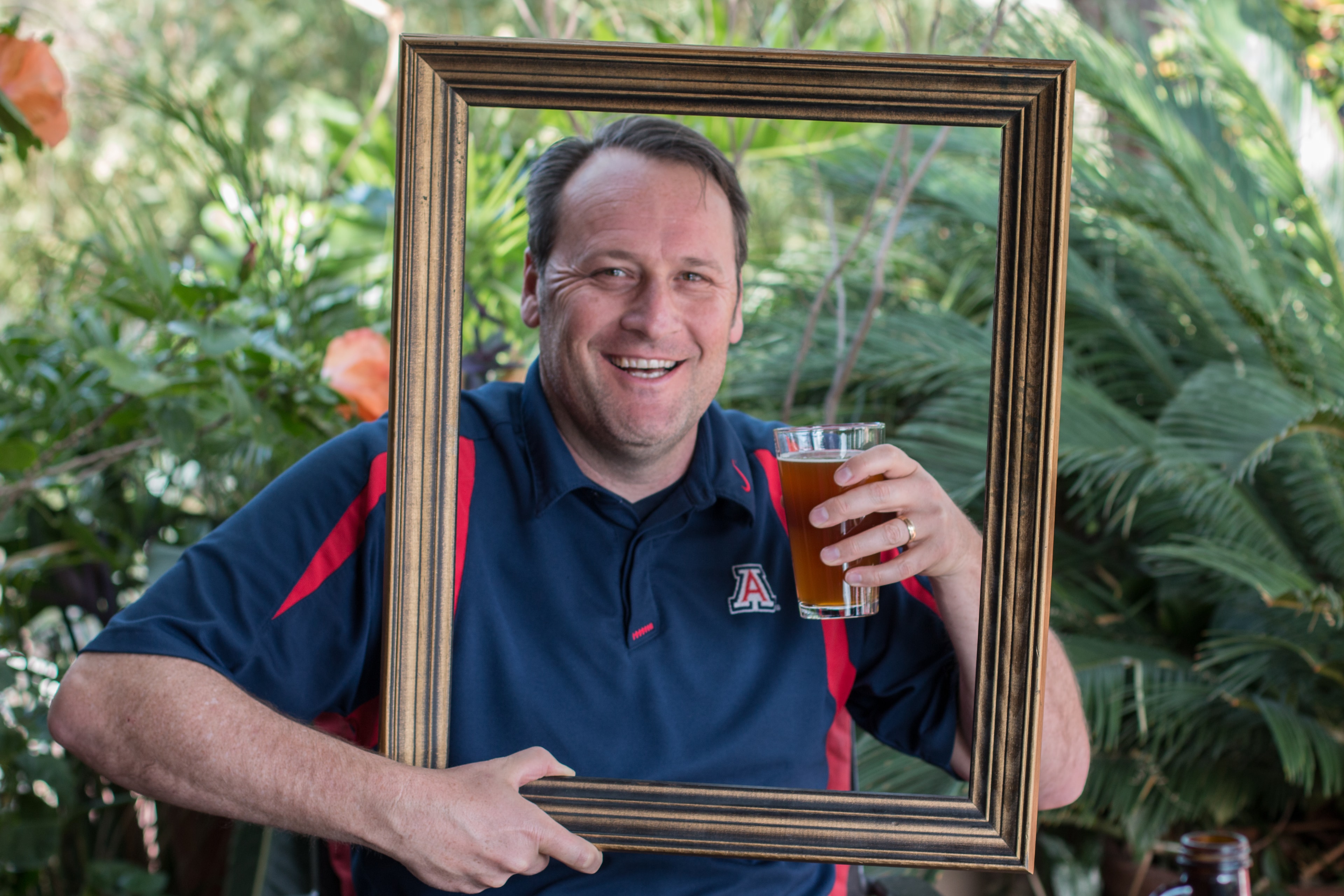 Smiling man in a University of Arizona polo holding a gold picture frame and a pint of beer in a lush garden