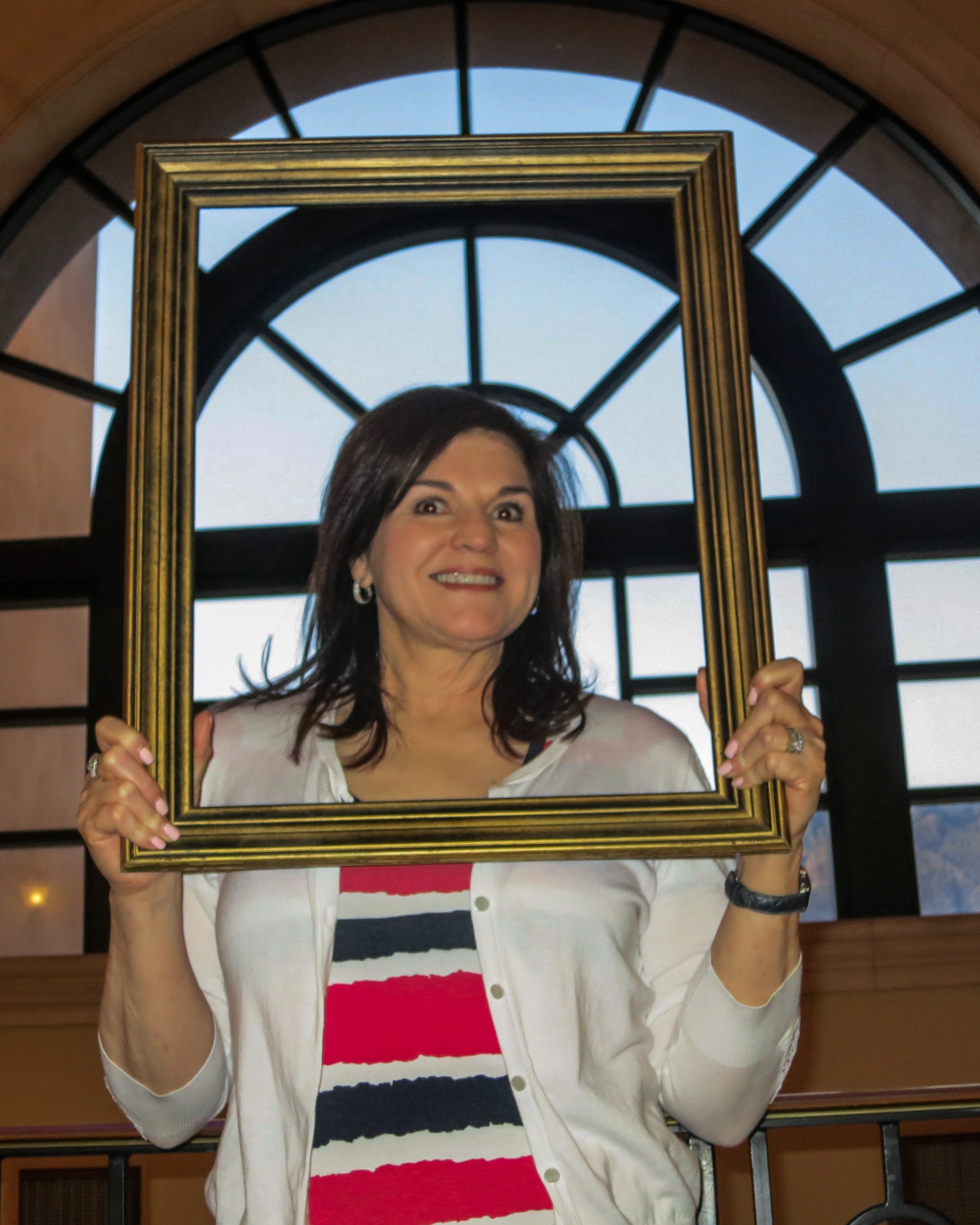 Woman holding a gold picture frame in front of a grand arched window with a striped cardigan and wide eyes