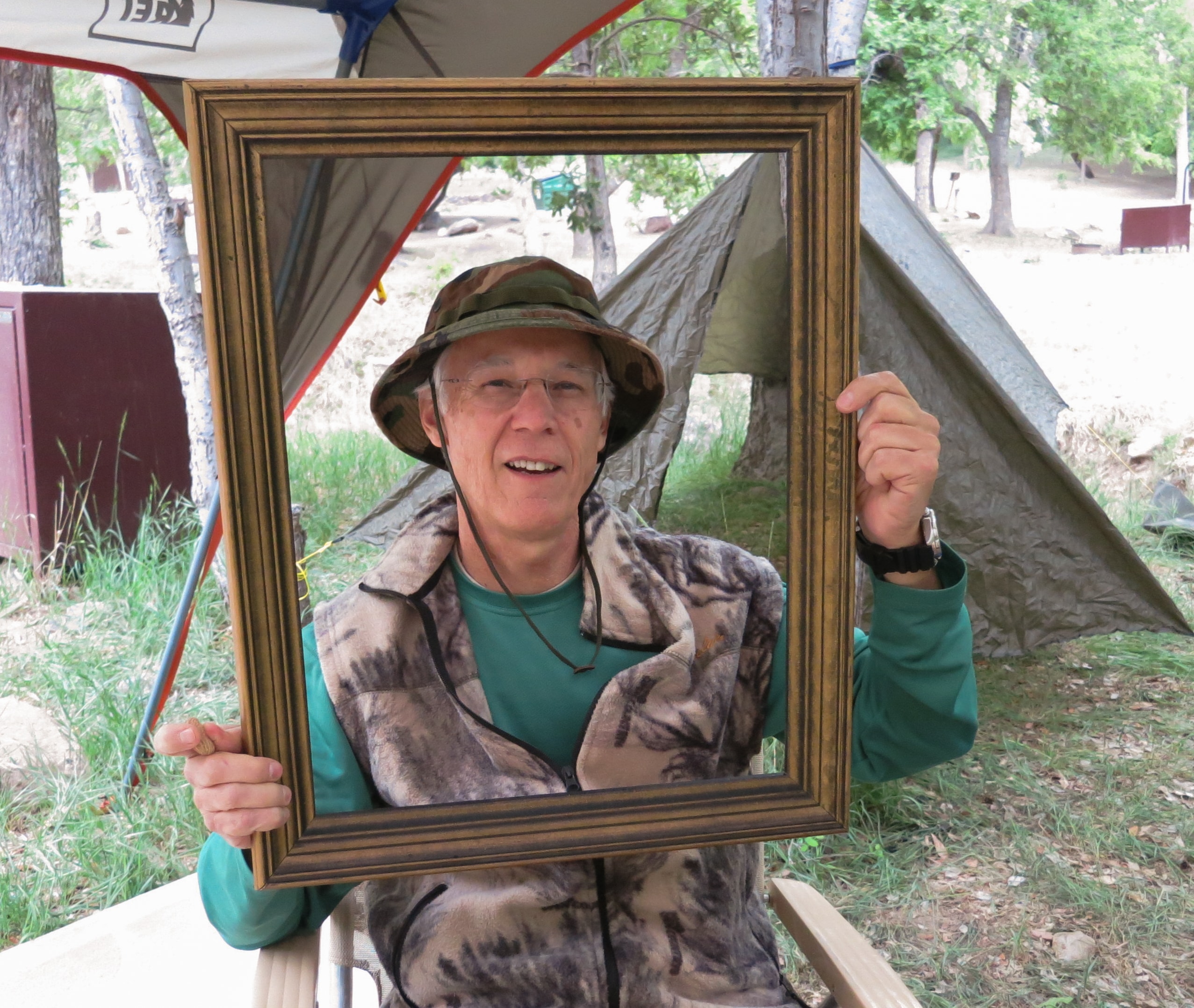 Man in a camo hat and fleece vest holding a gold picture frame at a campsite with tents in the background