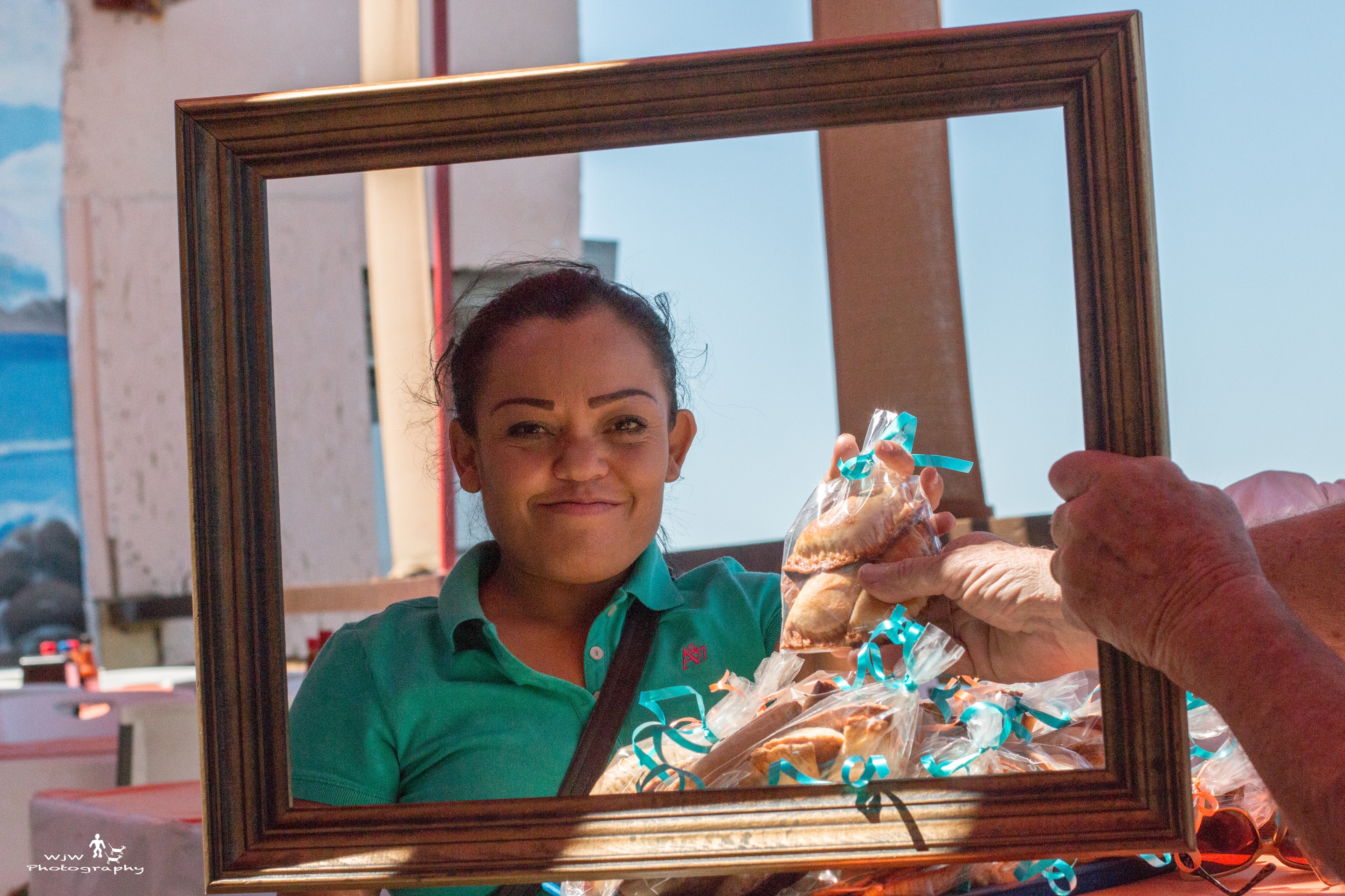 Woman in a teal polo smiling through a gold picture frame while holding packaged empanadas at an outdoor market