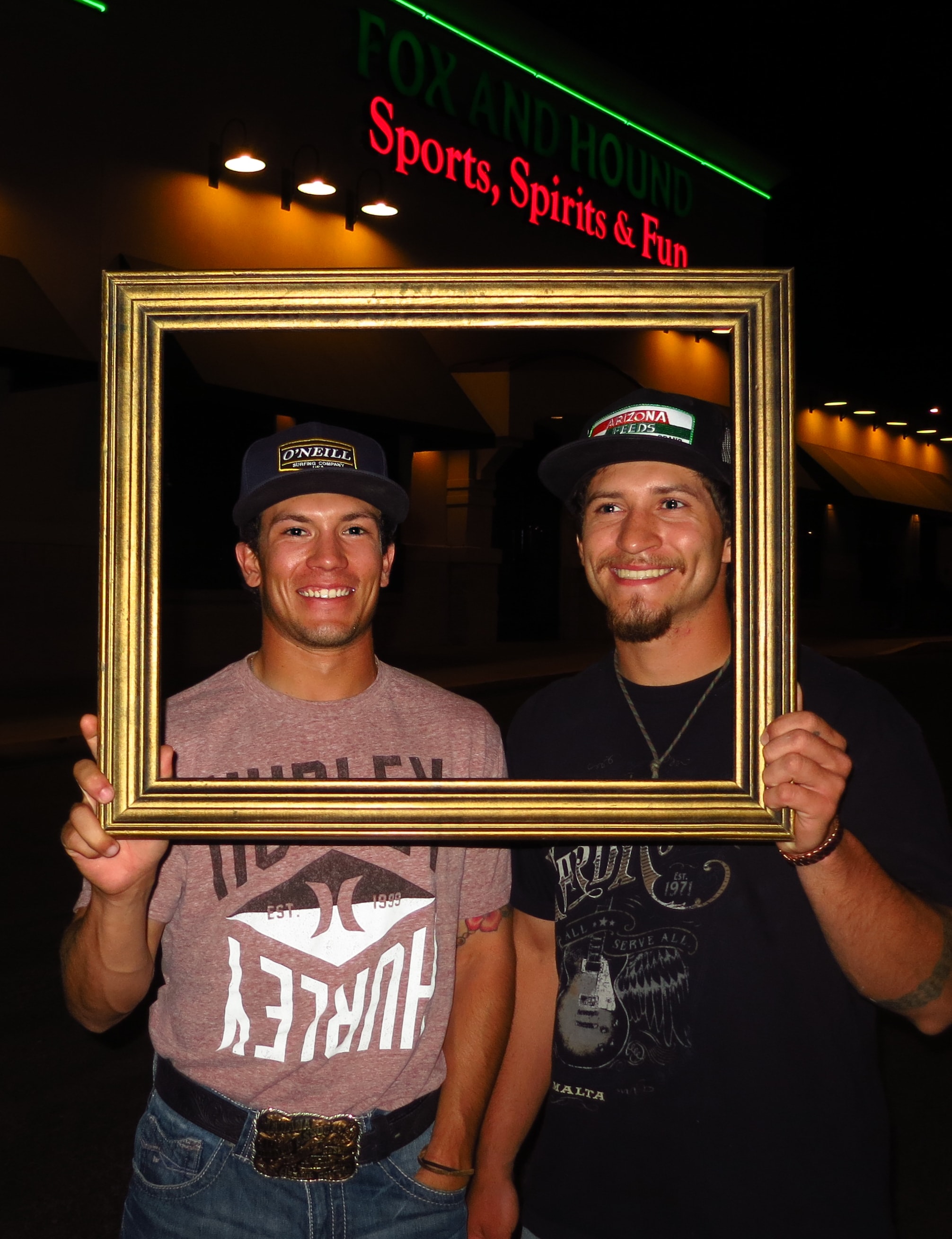Two young men in baseball caps holding a gold picture frame together outside the Fox and Hound sports bar at night