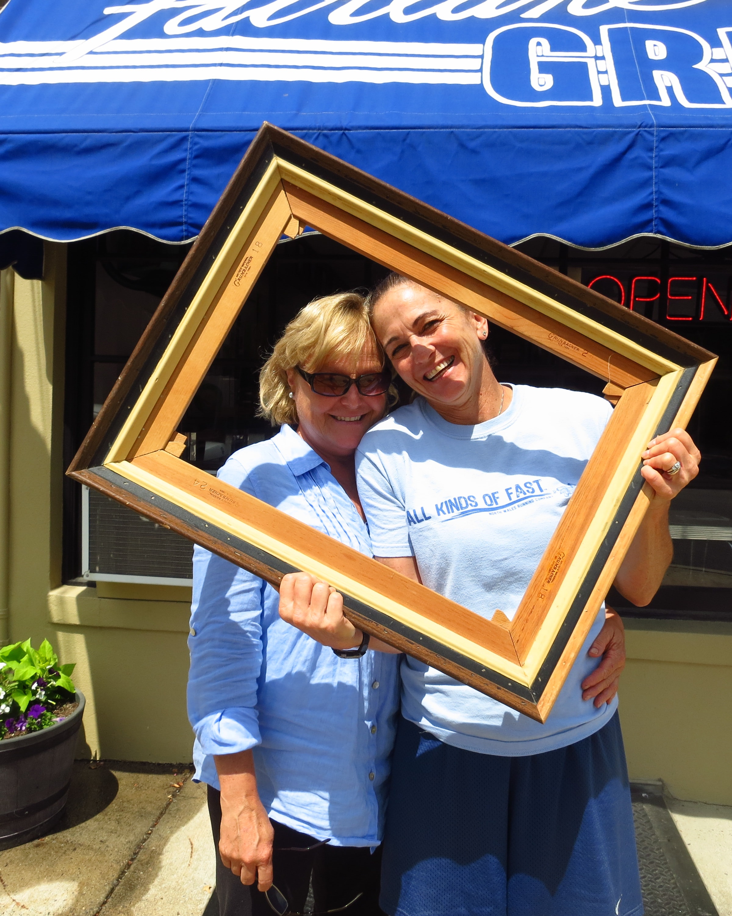 Two women laughing and leaning into a tilted wooden picture frame in front of a restaurant with a blue awning