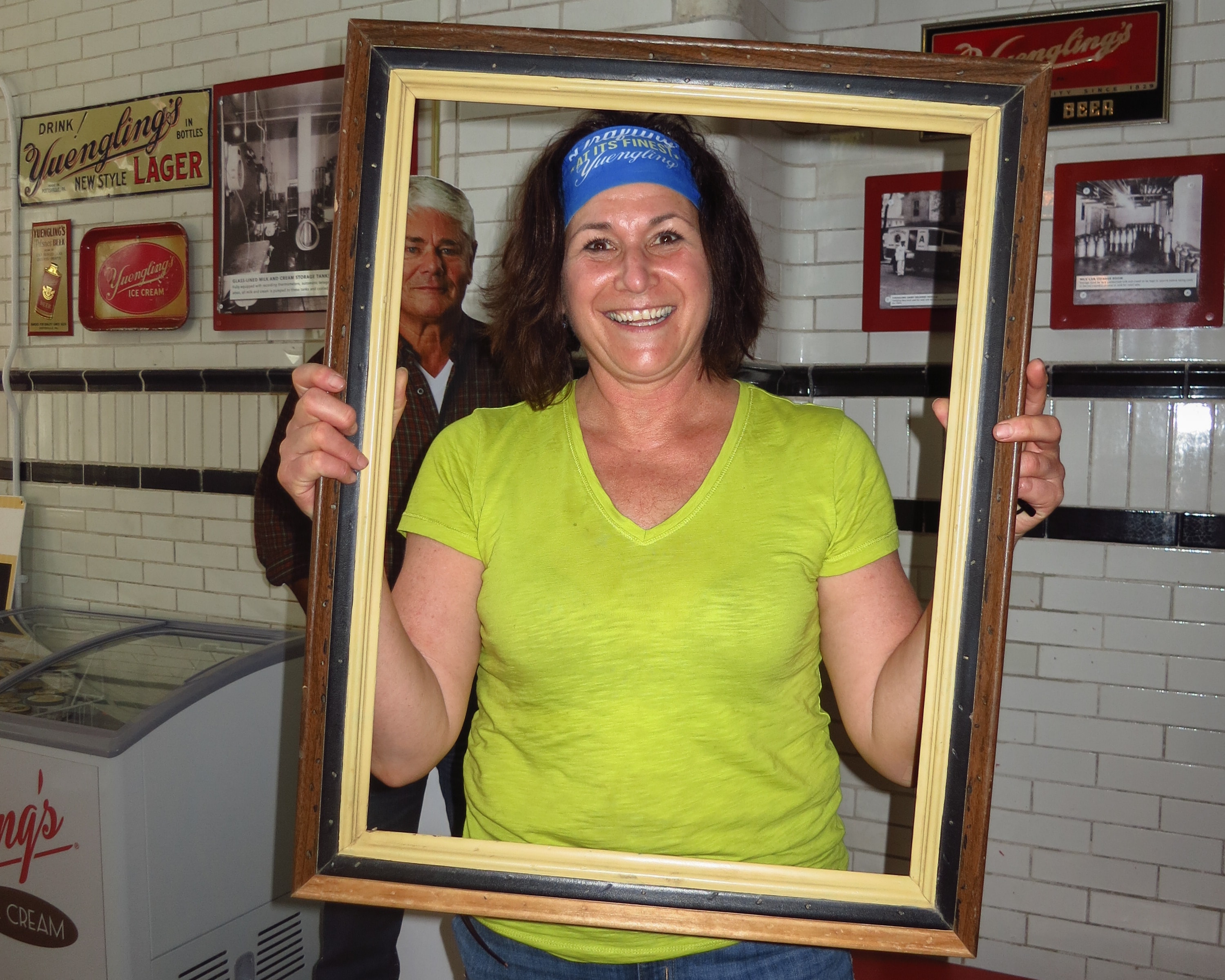 Woman in a lime green shirt and Yuengling headband holding a picture frame inside the Yuengling brewery with vintage signage on the walls