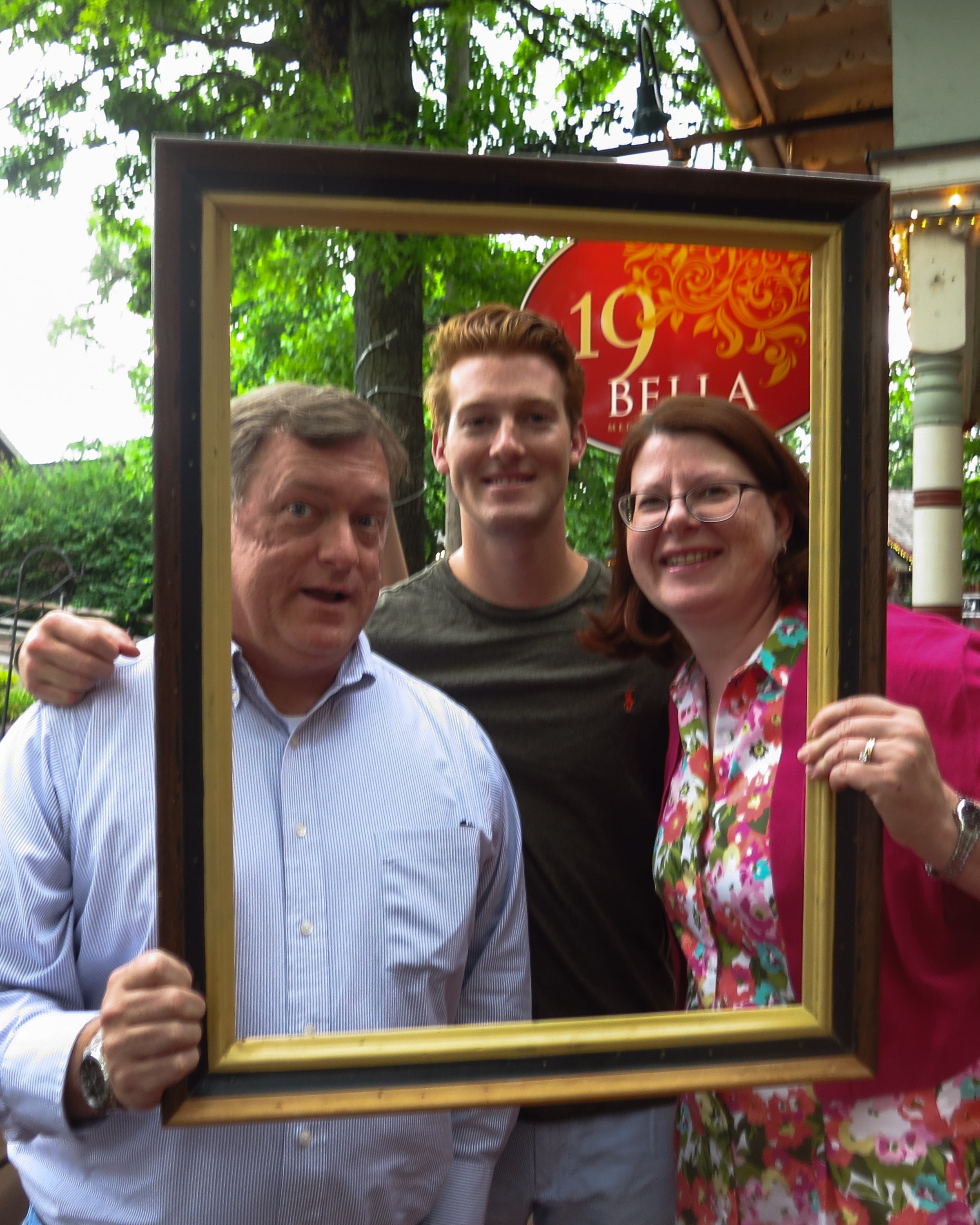 Three people — two men and a woman — crowding into a picture frame on the patio of Bella restaurant with lush green trees behind them