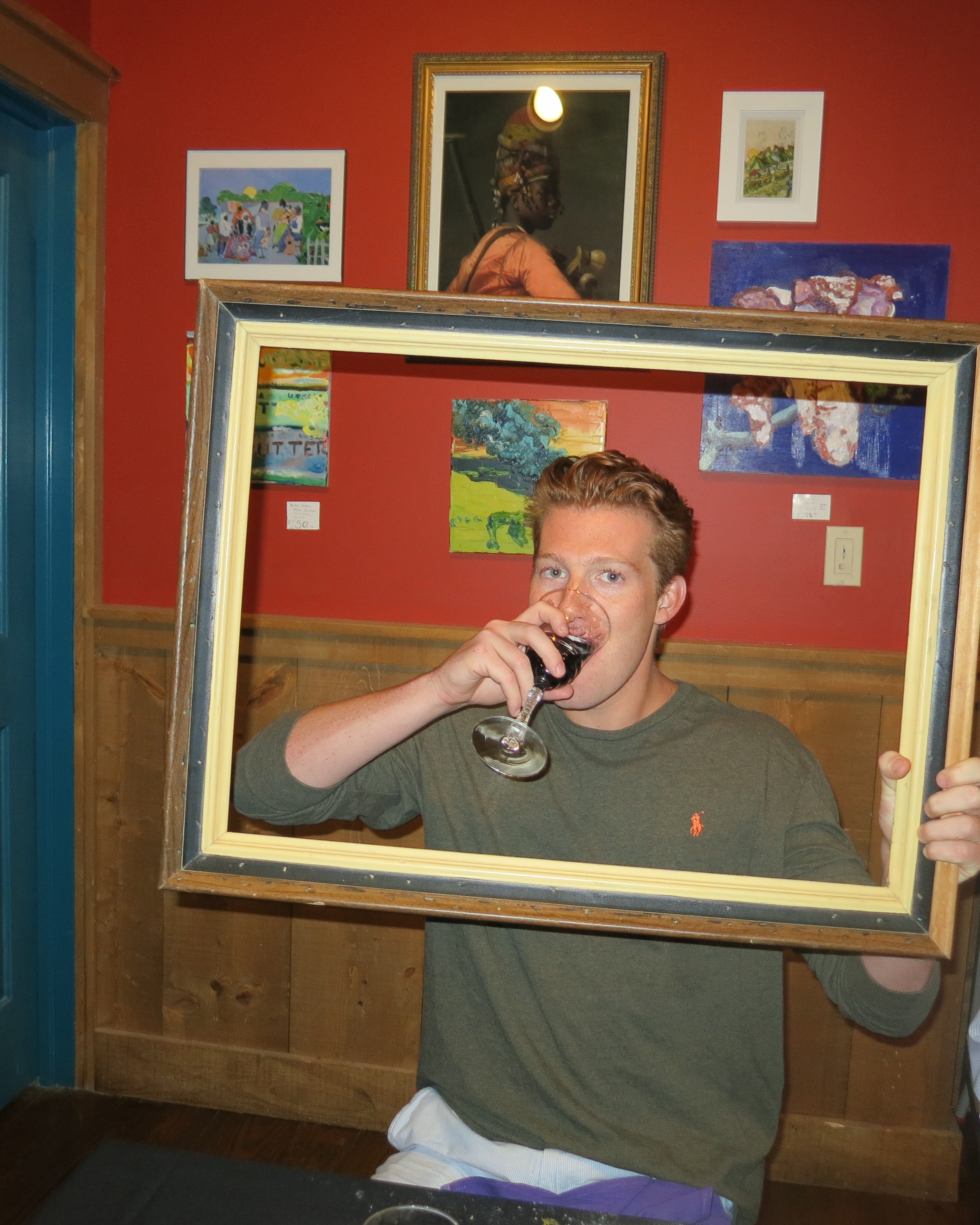 Young man sipping red wine through a picture frame inside an art gallery with red walls and paintings behind him
