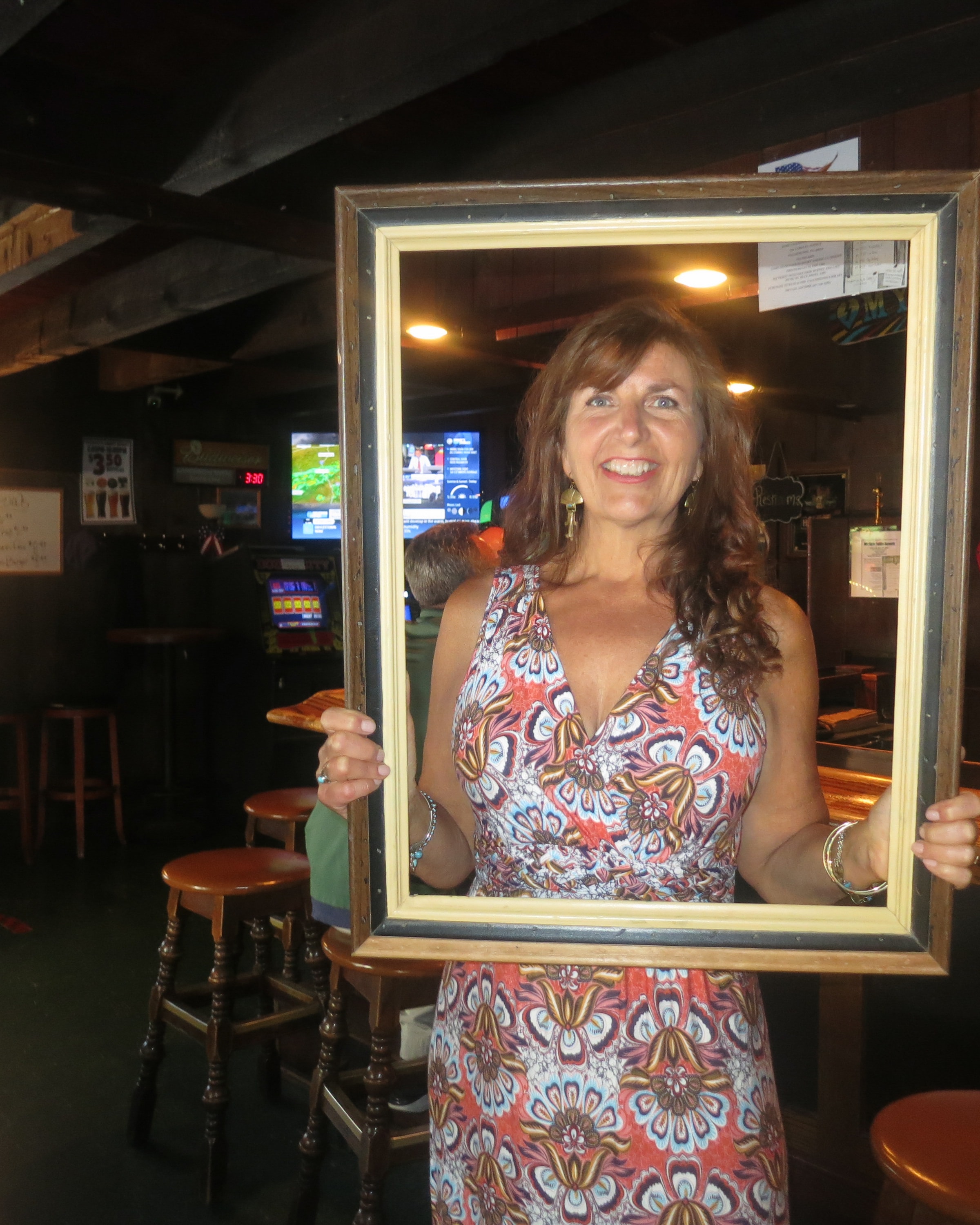 Woman in a colorful floral dress holding a picture frame and smiling inside a dimly lit bar with TVs and bar stools behind her