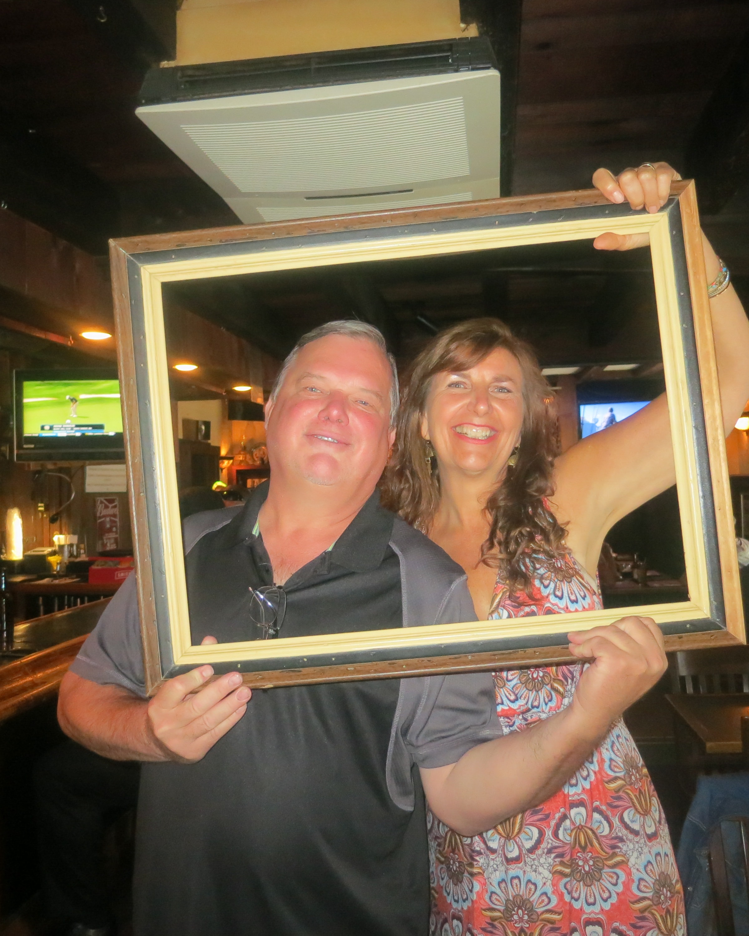 Man and woman smiling together inside a picture frame at a bar, TVs and warm bar lighting glowing behind them
