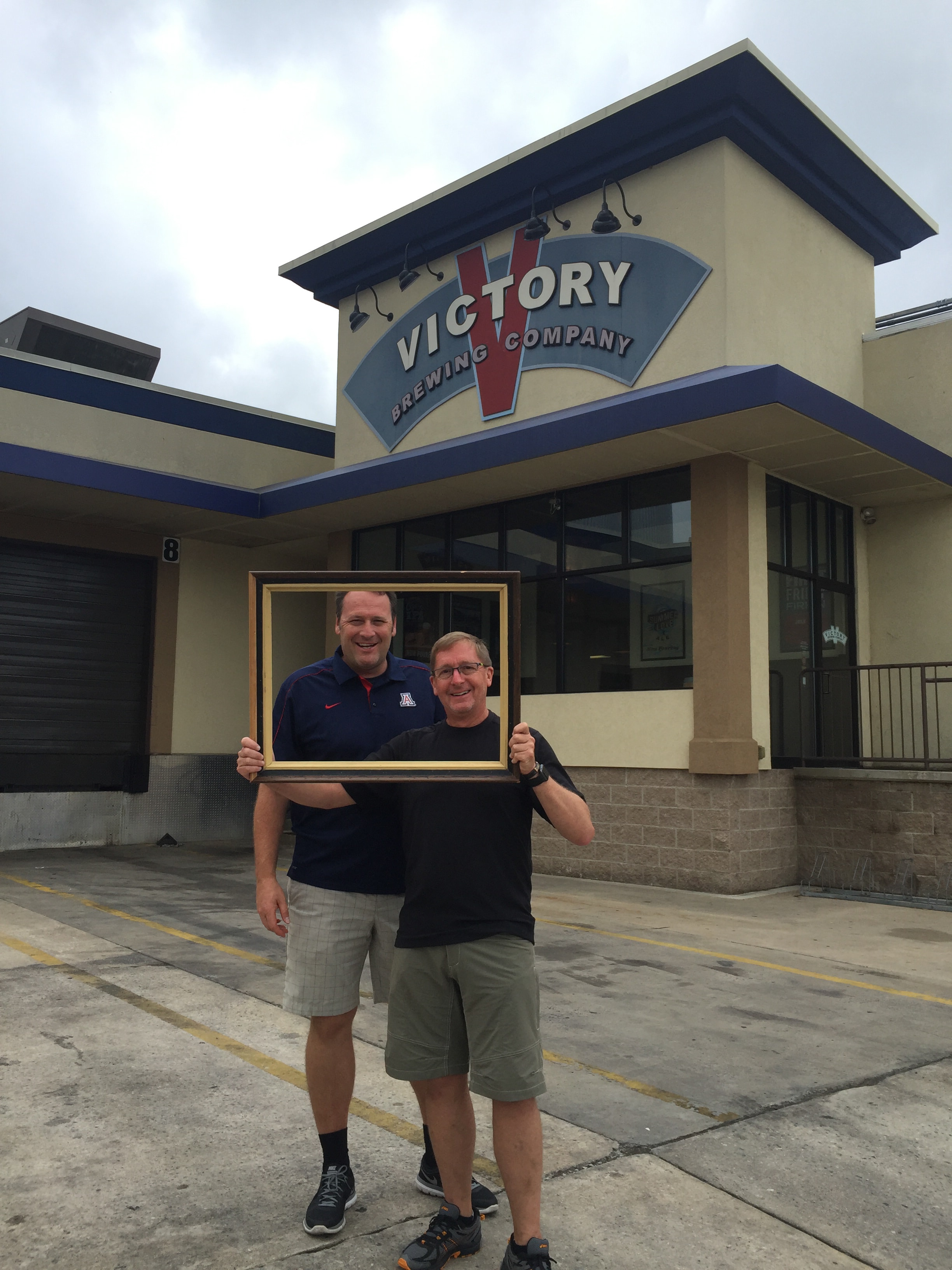 Two men holding a gold picture frame together in front of the Victory Brewing Company sign
