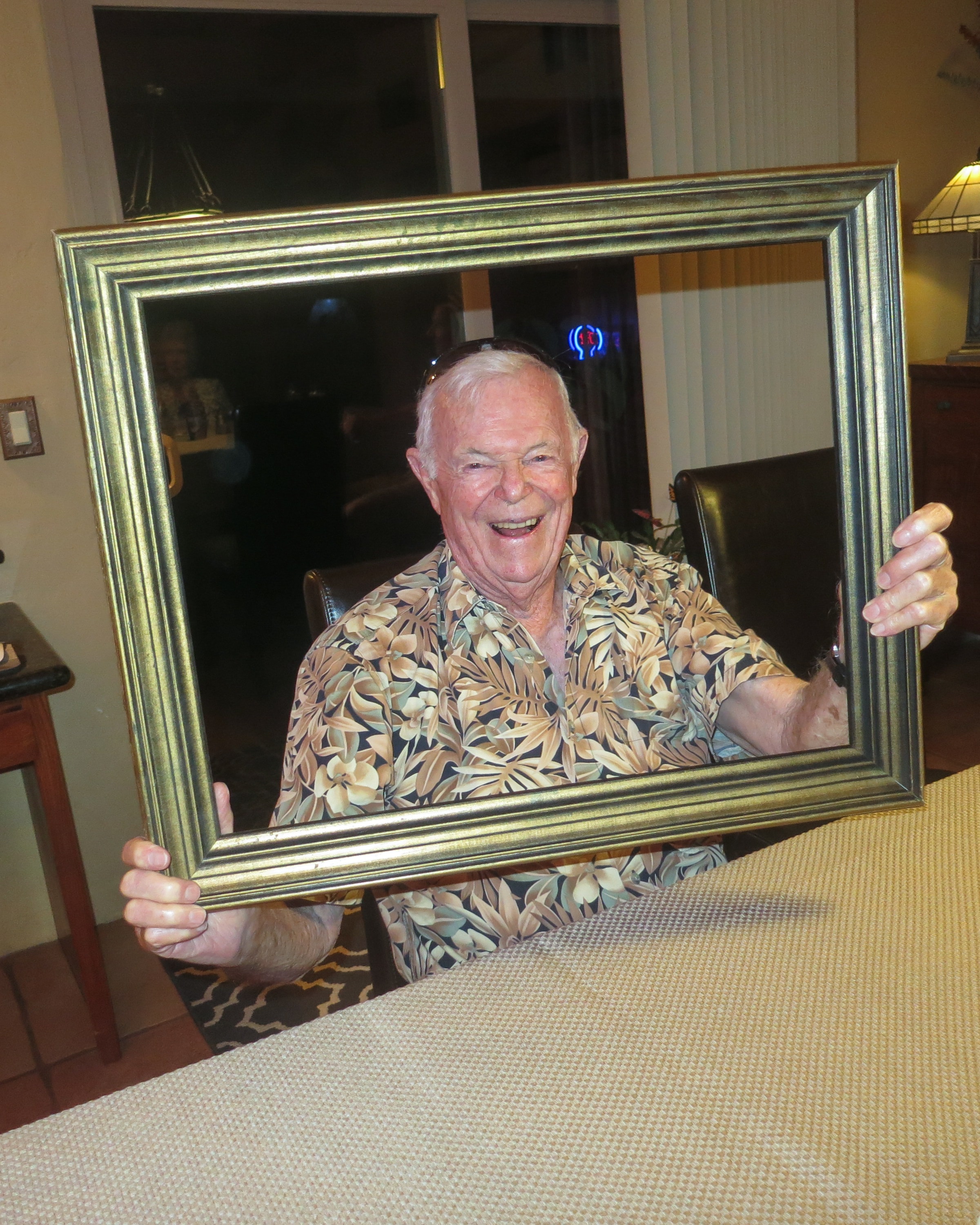 Elderly man in a Hawaiian shirt laughing broadly while holding a large gold picture frame at a dining table