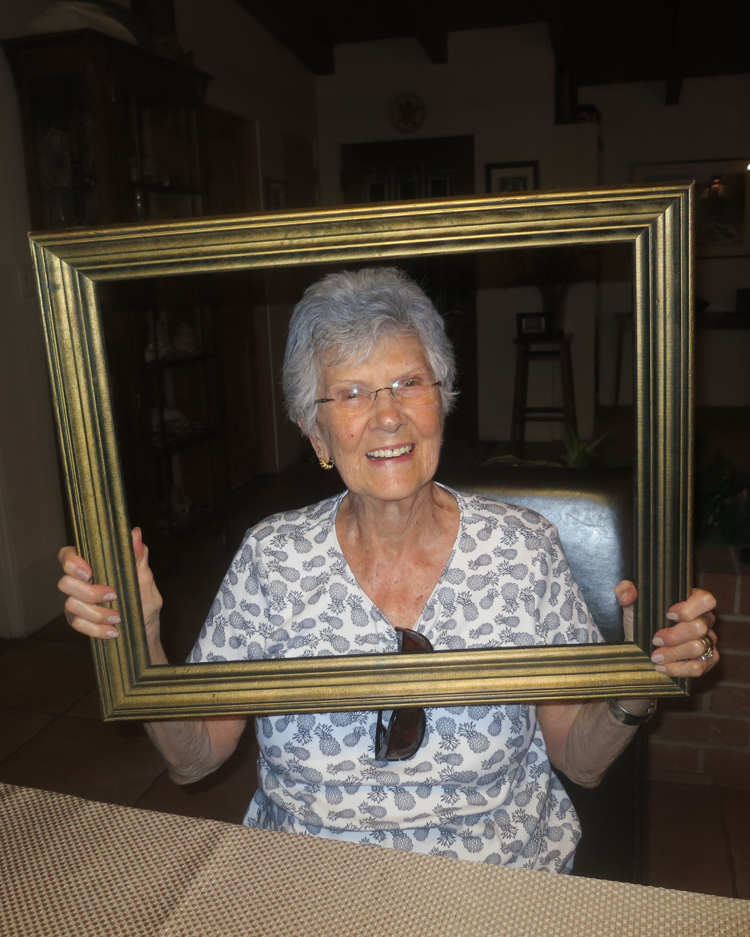 Elderly woman with silver hair and a pineapple-print blouse smiling warmly through a large gold picture frame