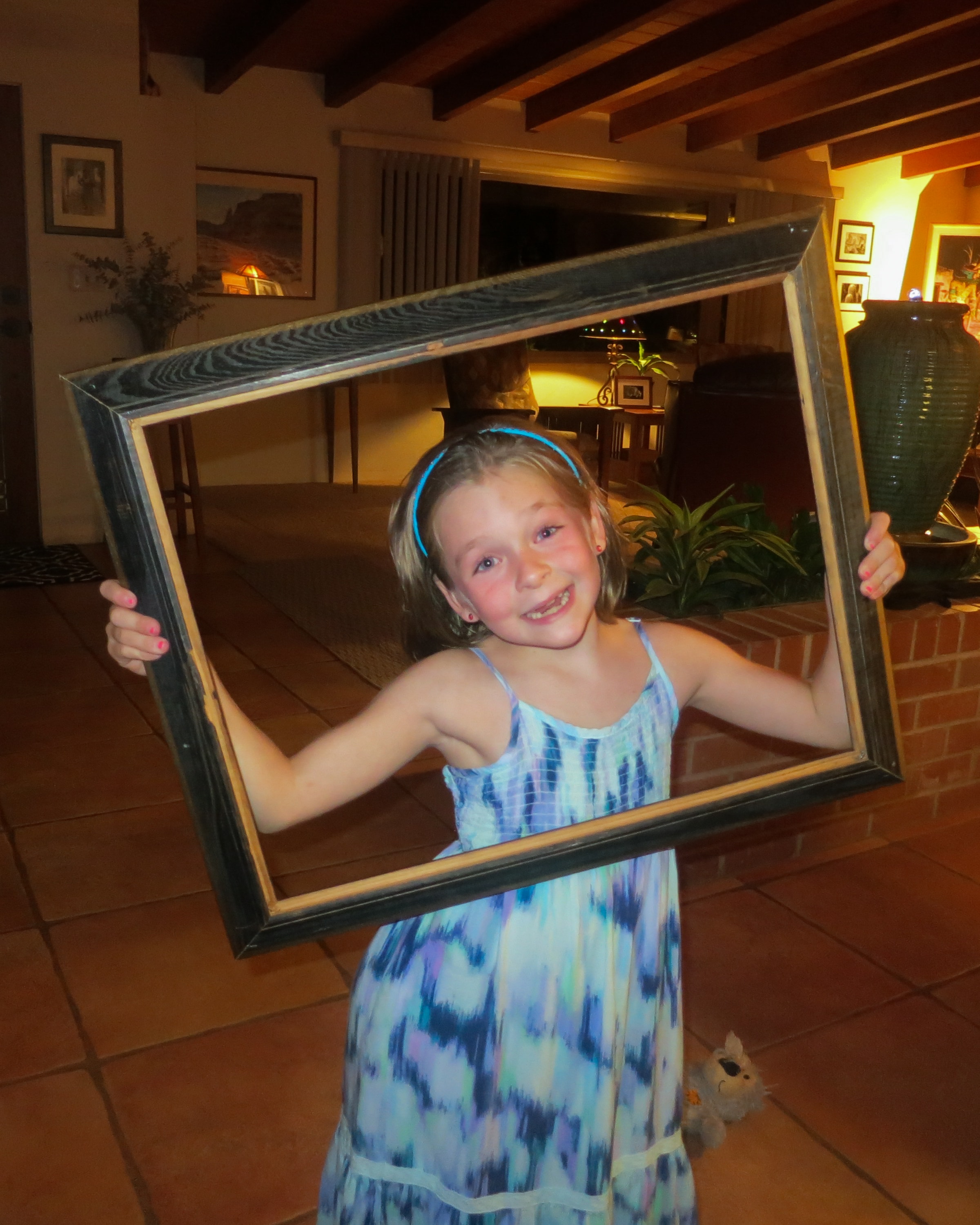 Young girl in a tie-dye dress and blue headband holding a large dark wooden picture frame indoors with warm lighting