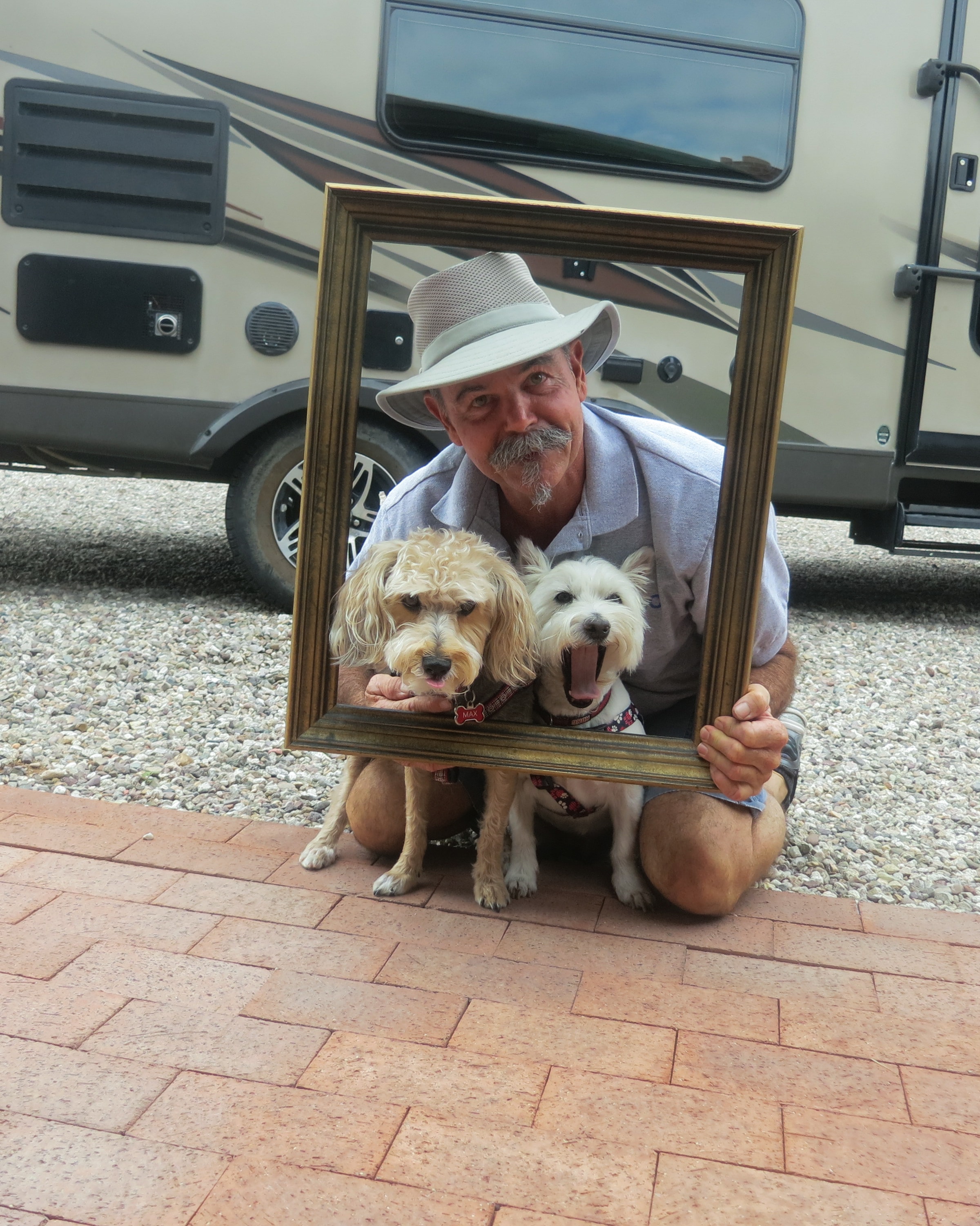 Man in a straw hat crouching with two small dogs — a goldendoodle and a westie — all framed together in a gold picture frame in front of an RV