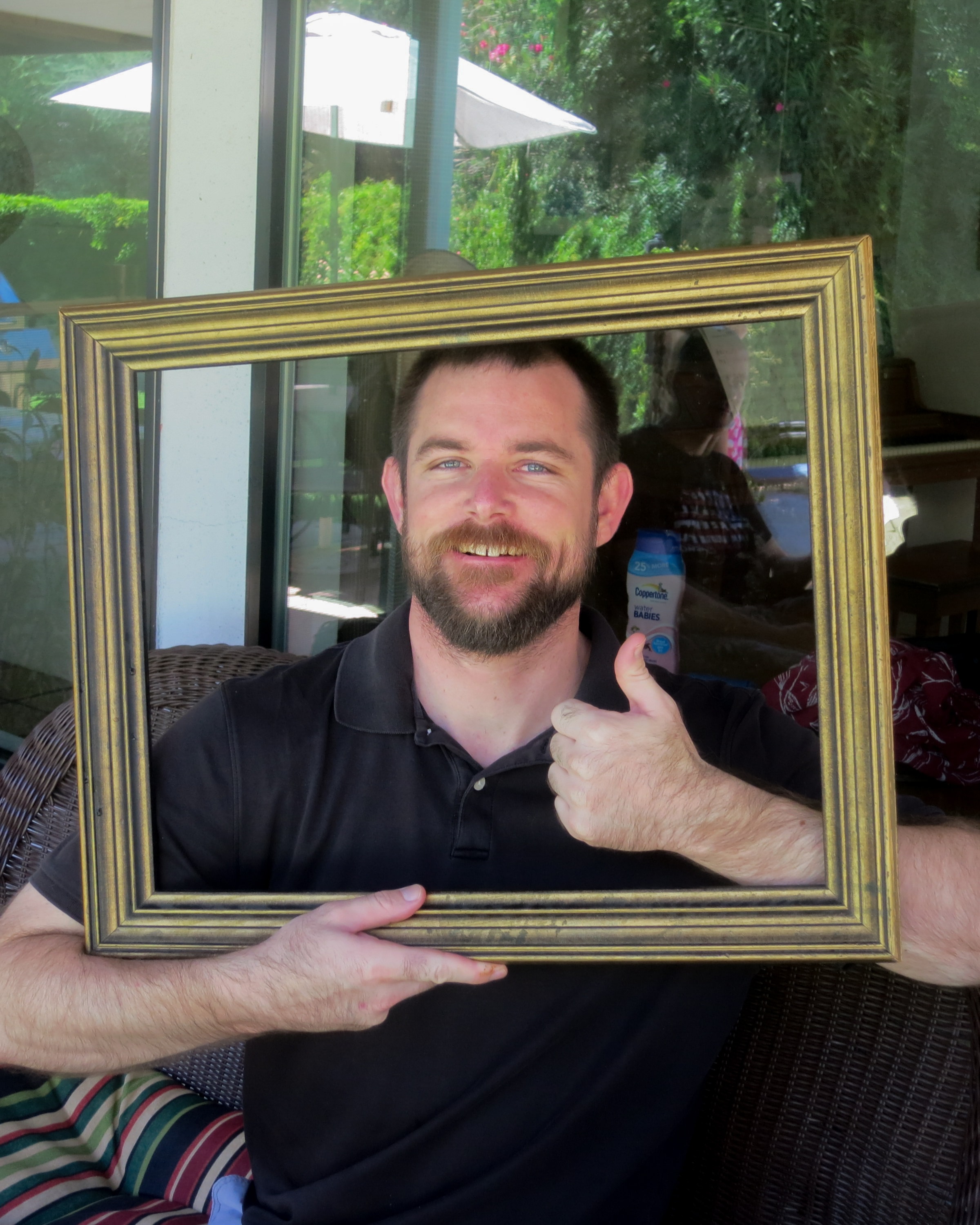 Bearded man in a black polo giving a thumbs up through a gold picture frame on a shaded patio with lush greenery behind him
