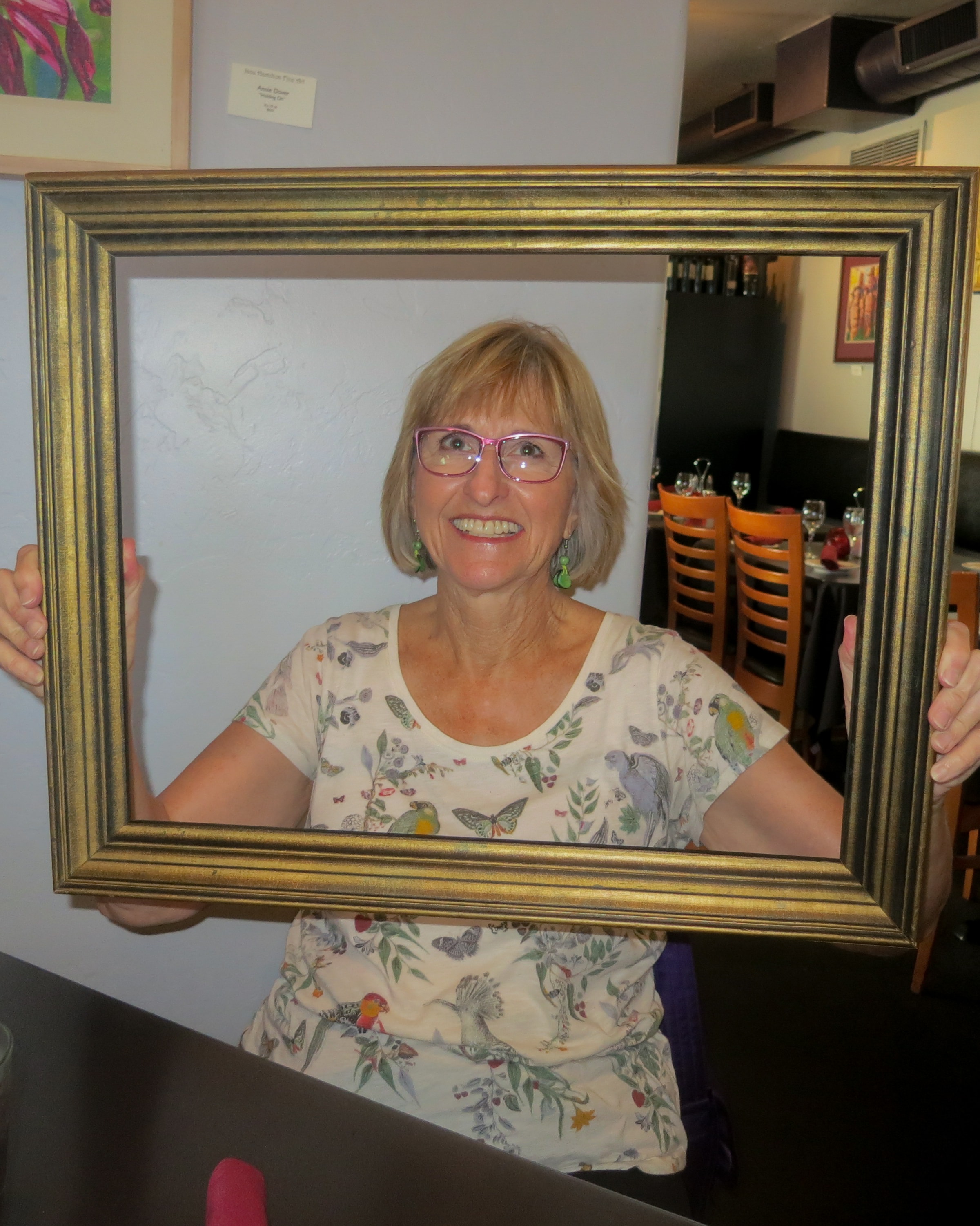 Woman with pink glasses and a bird-print blouse beaming through a gold picture frame inside a restaurant with artwork on the walls