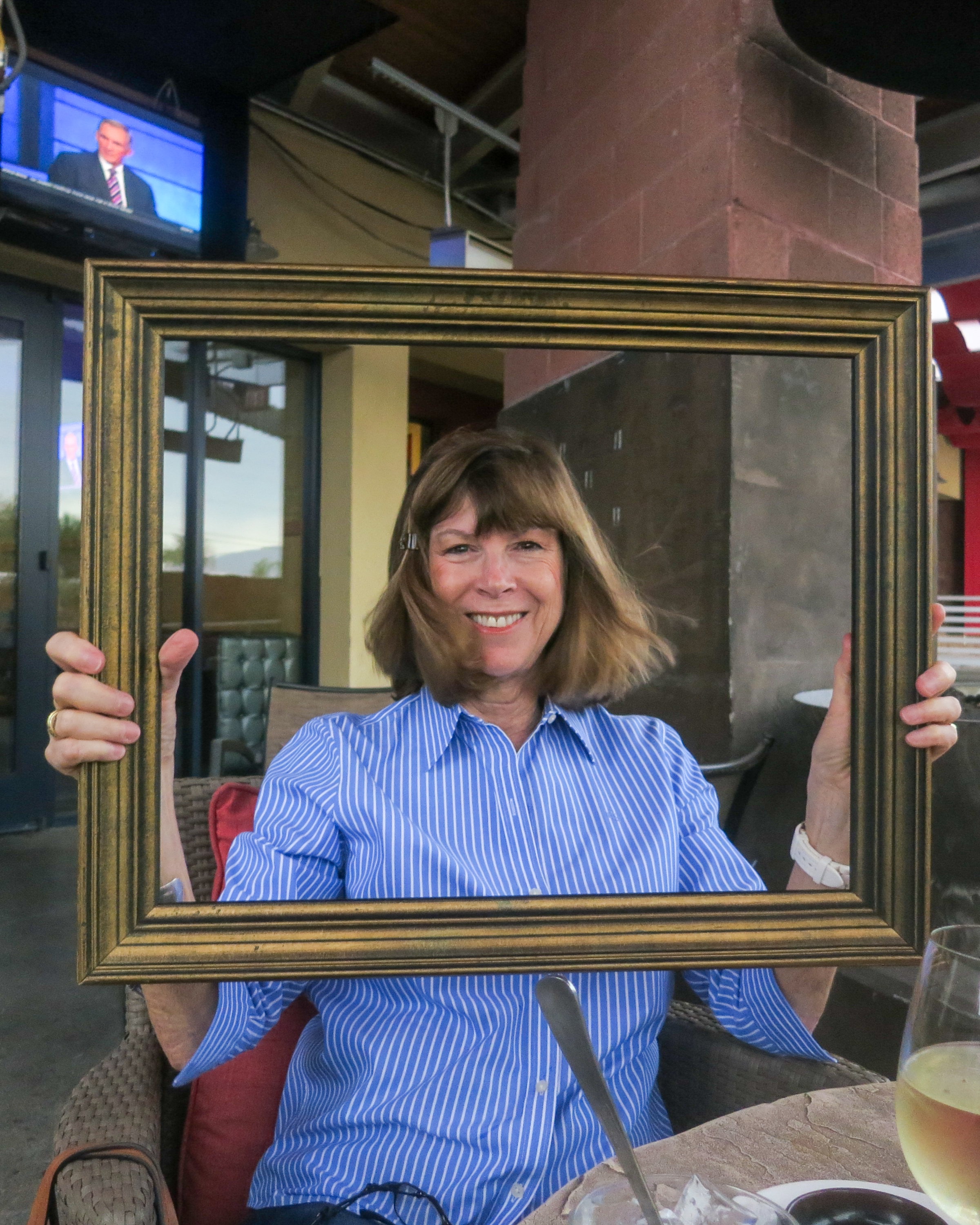 Woman in a blue striped shirt smiling through a gold picture frame on a restaurant patio with a TV and fireplace behind her