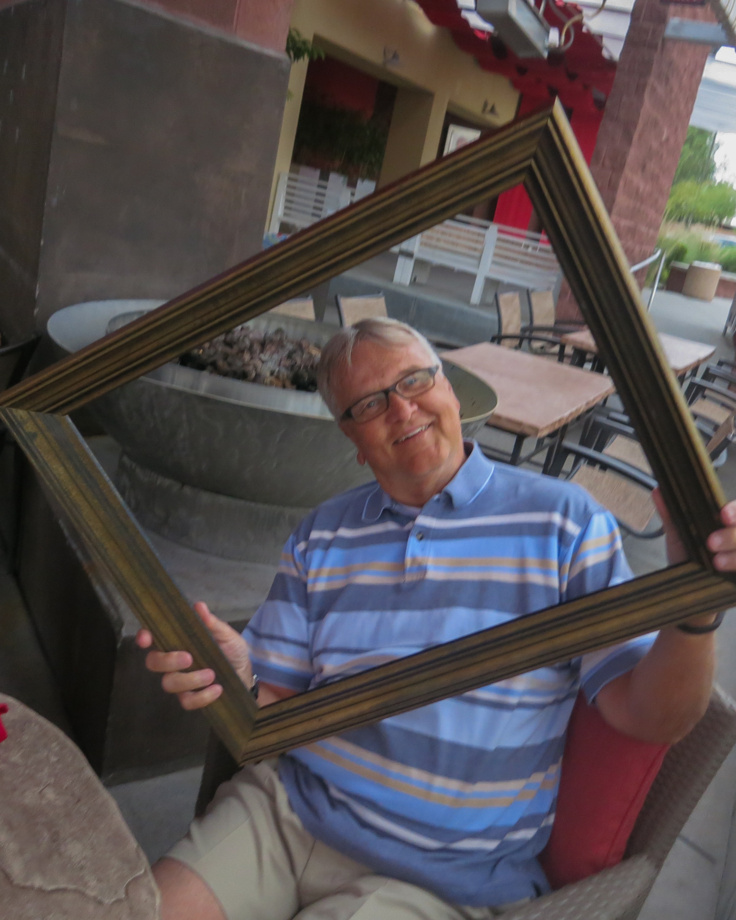 Man in a striped polo holding a tilted gold picture frame on a restaurant patio with a fire pit and outdoor seating behind him