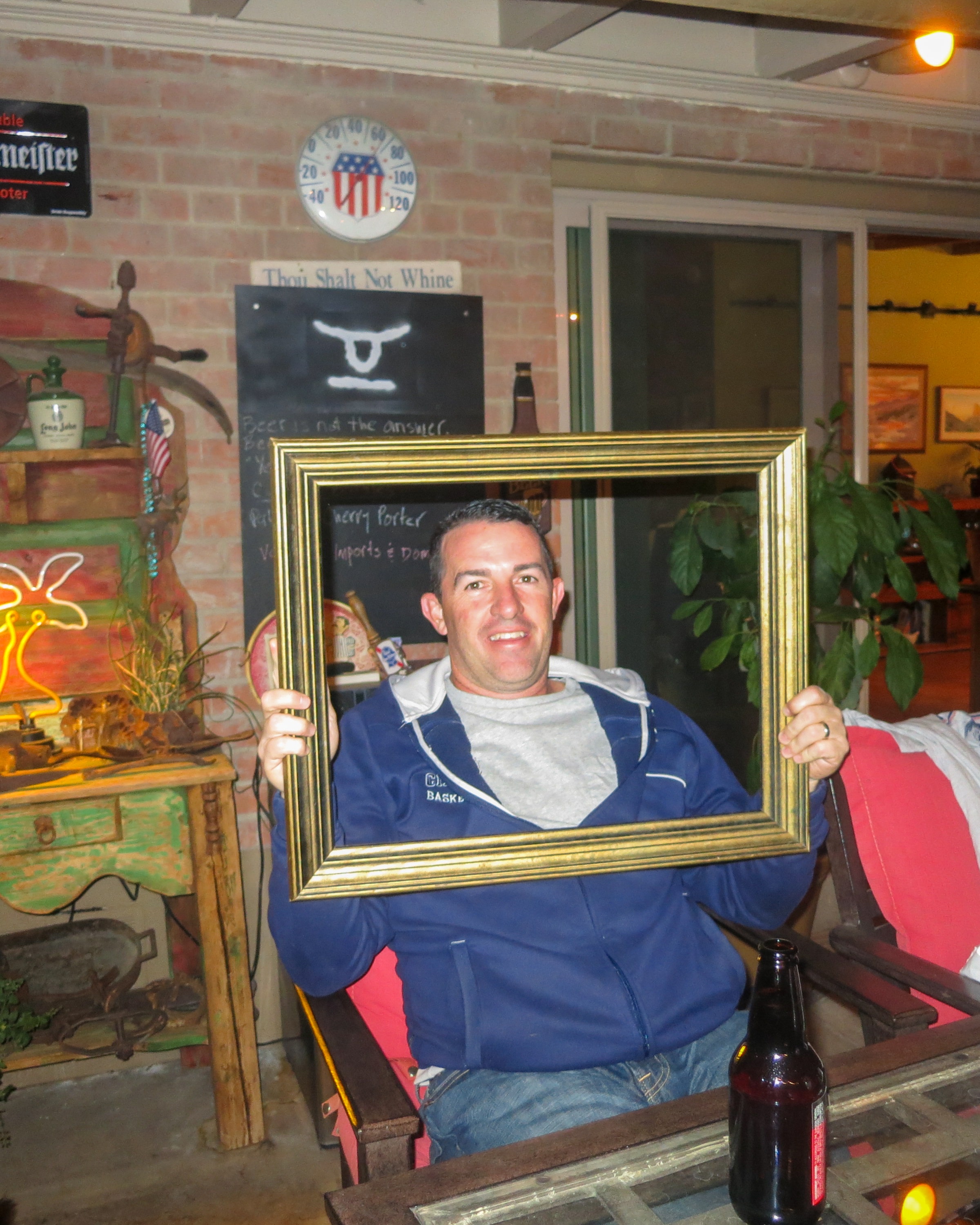 Man in a blue basketball jacket holding a gold picture frame at a rustic bar with a chalkboard sign reading Thou Shalt Not Whine and a growler of beer beside him