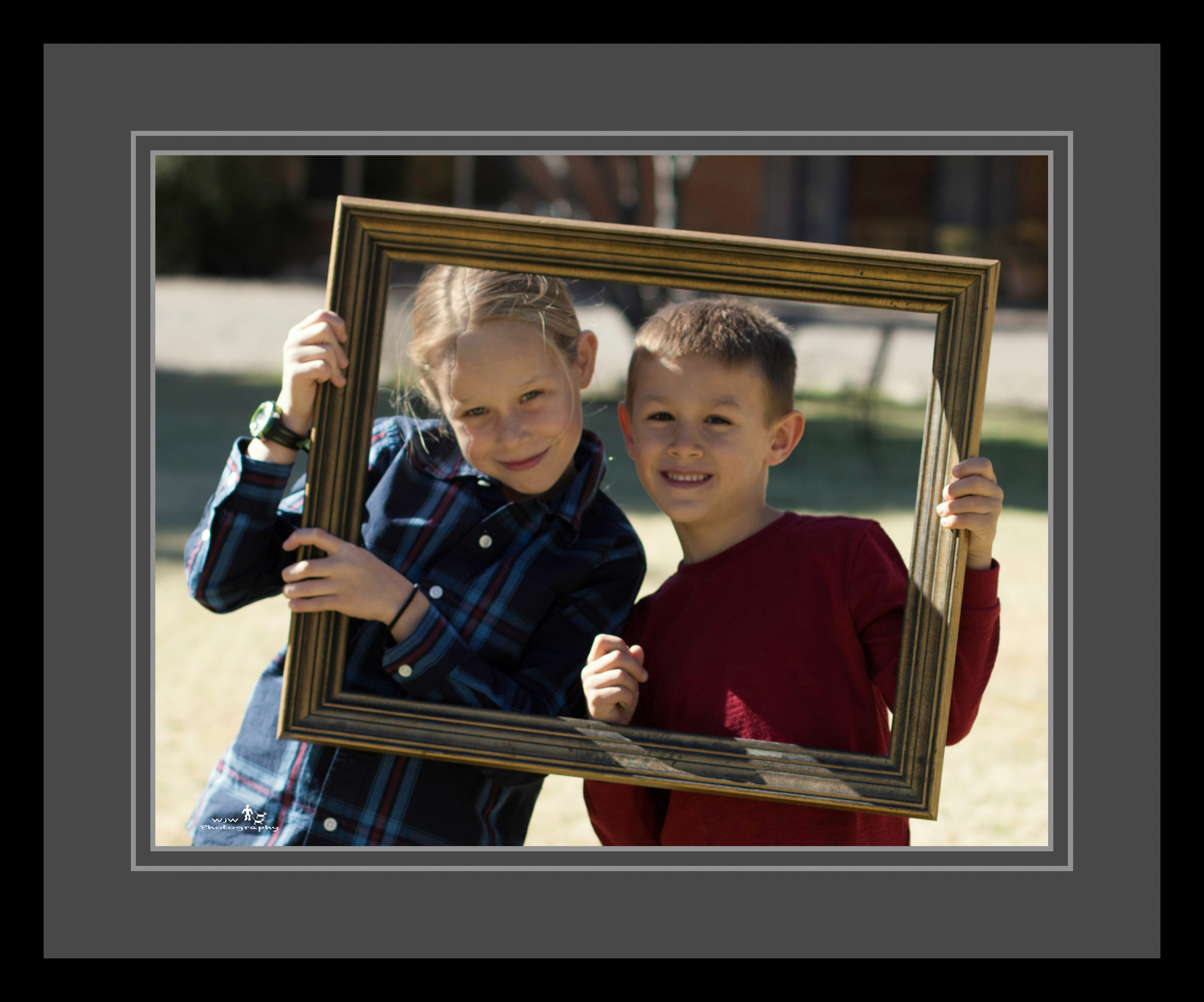 Two kids — a girl in a plaid shirt and a boy in red — grinning together through a gold picture frame outdoors in bright sunlight