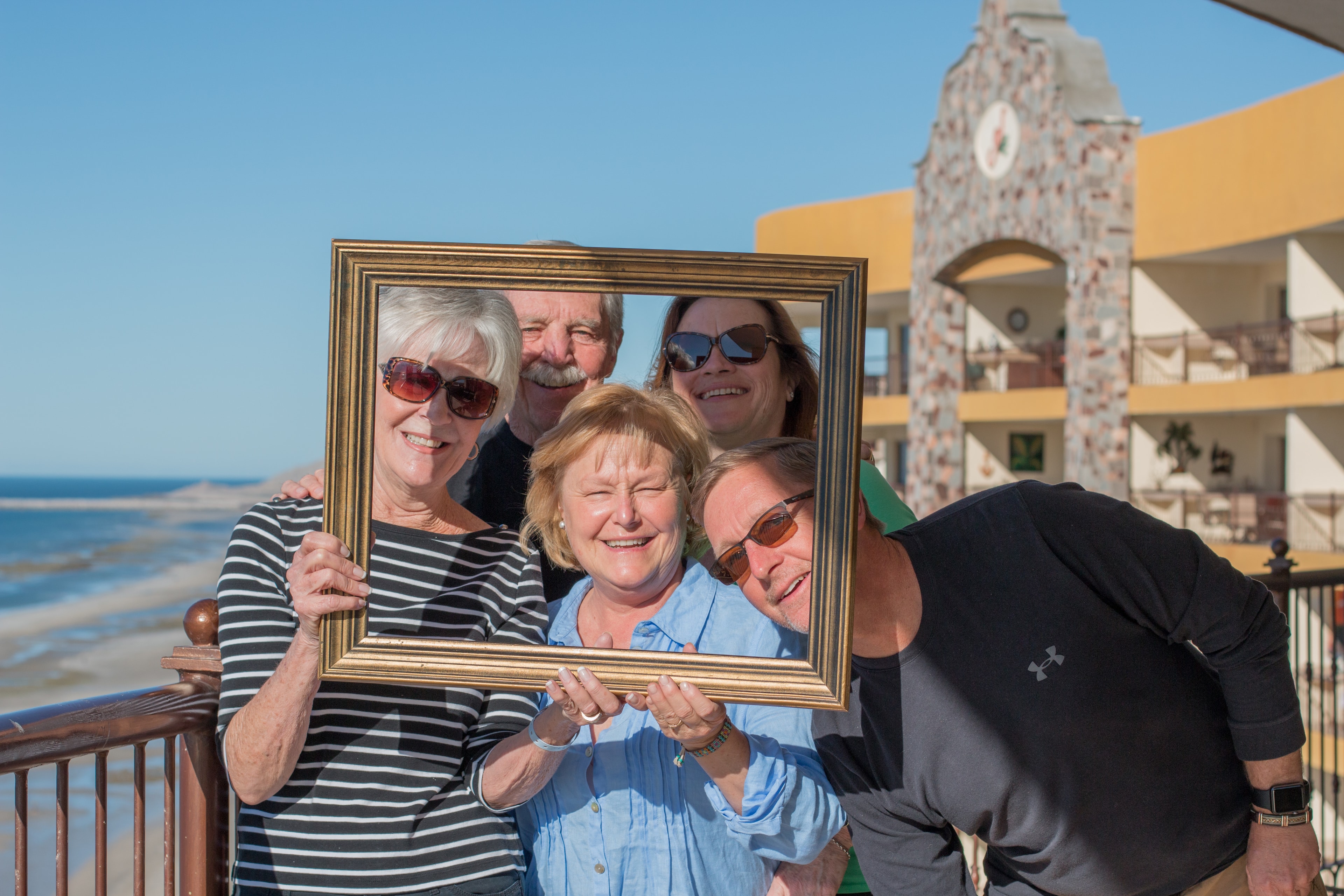 Five adults crowding together through a gold picture frame on a beachside terrace with the Sea of Cortez and a resort tower behind them