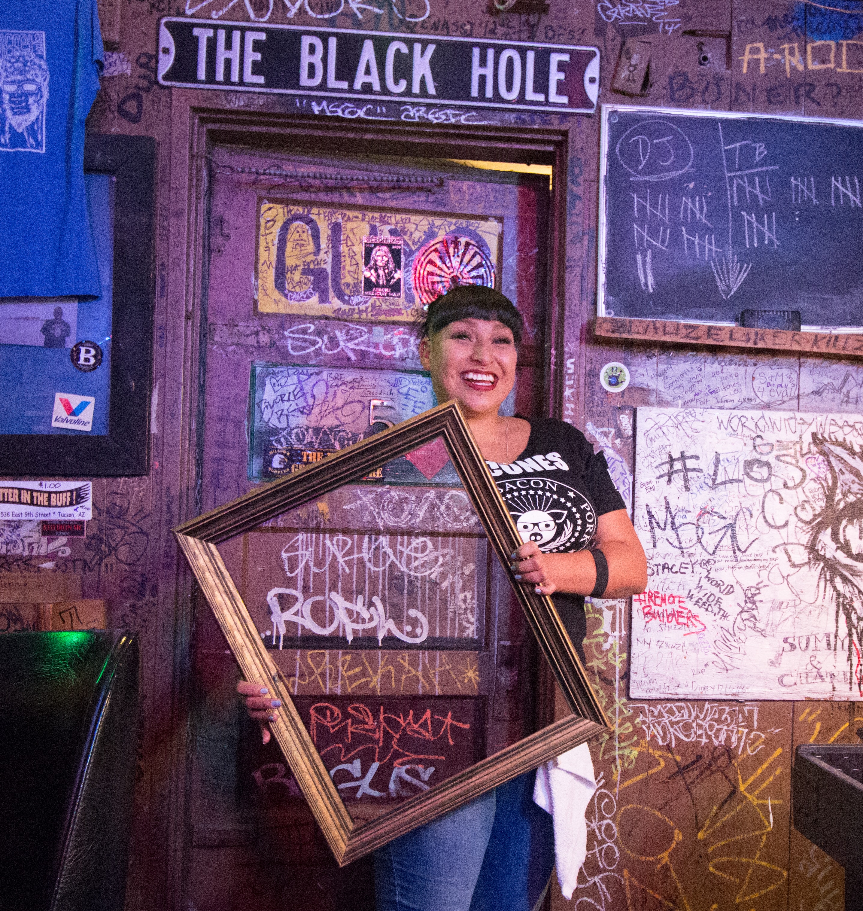 Young woman in a black band tee laughing while holding a large tilted picture frame inside The Black Hole bar, graffiti-covered walls surrounding her
