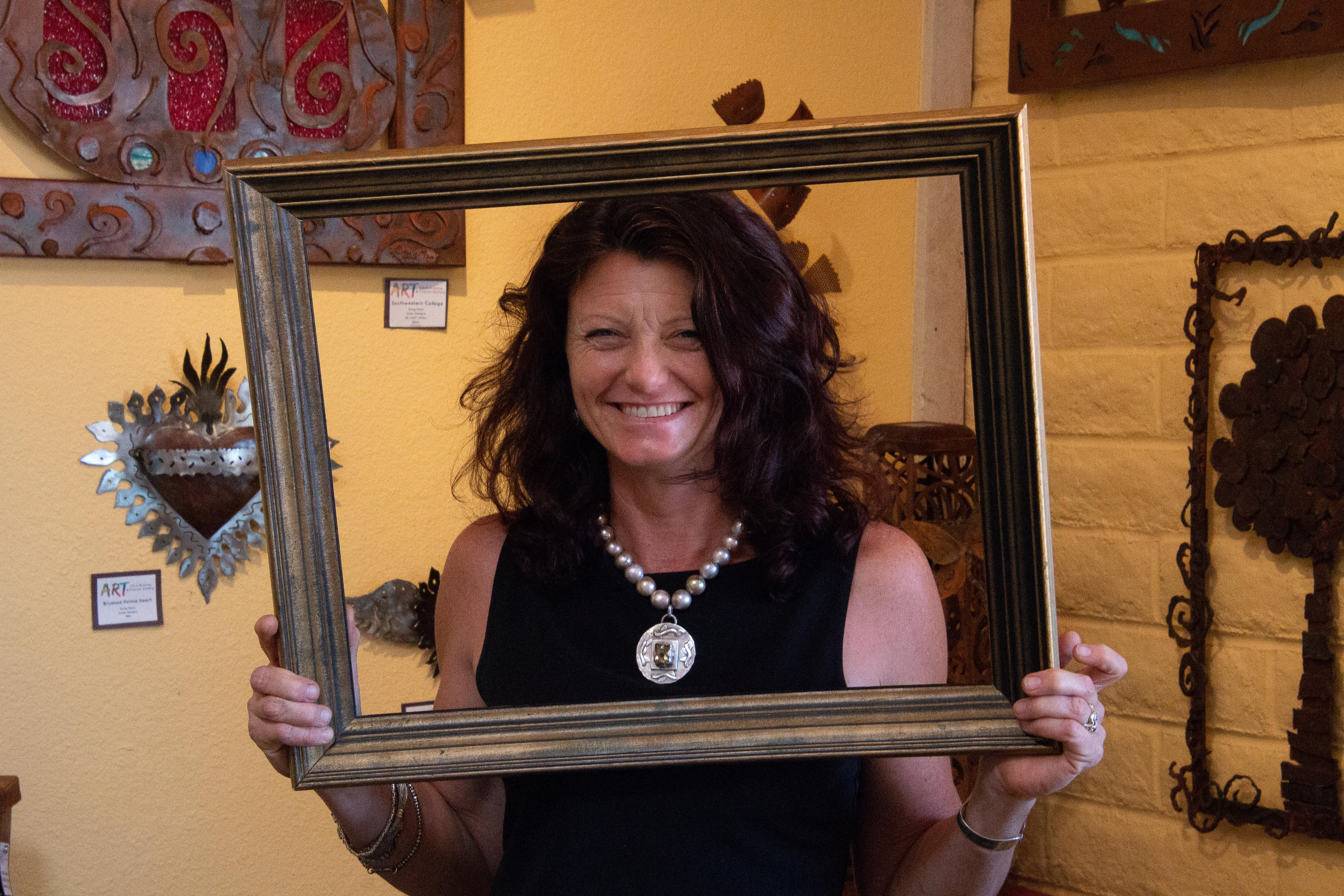 Woman with dark curly hair and a pearl necklace smiling through a large gold picture frame inside a gallery filled with ornate metal wall sculptures