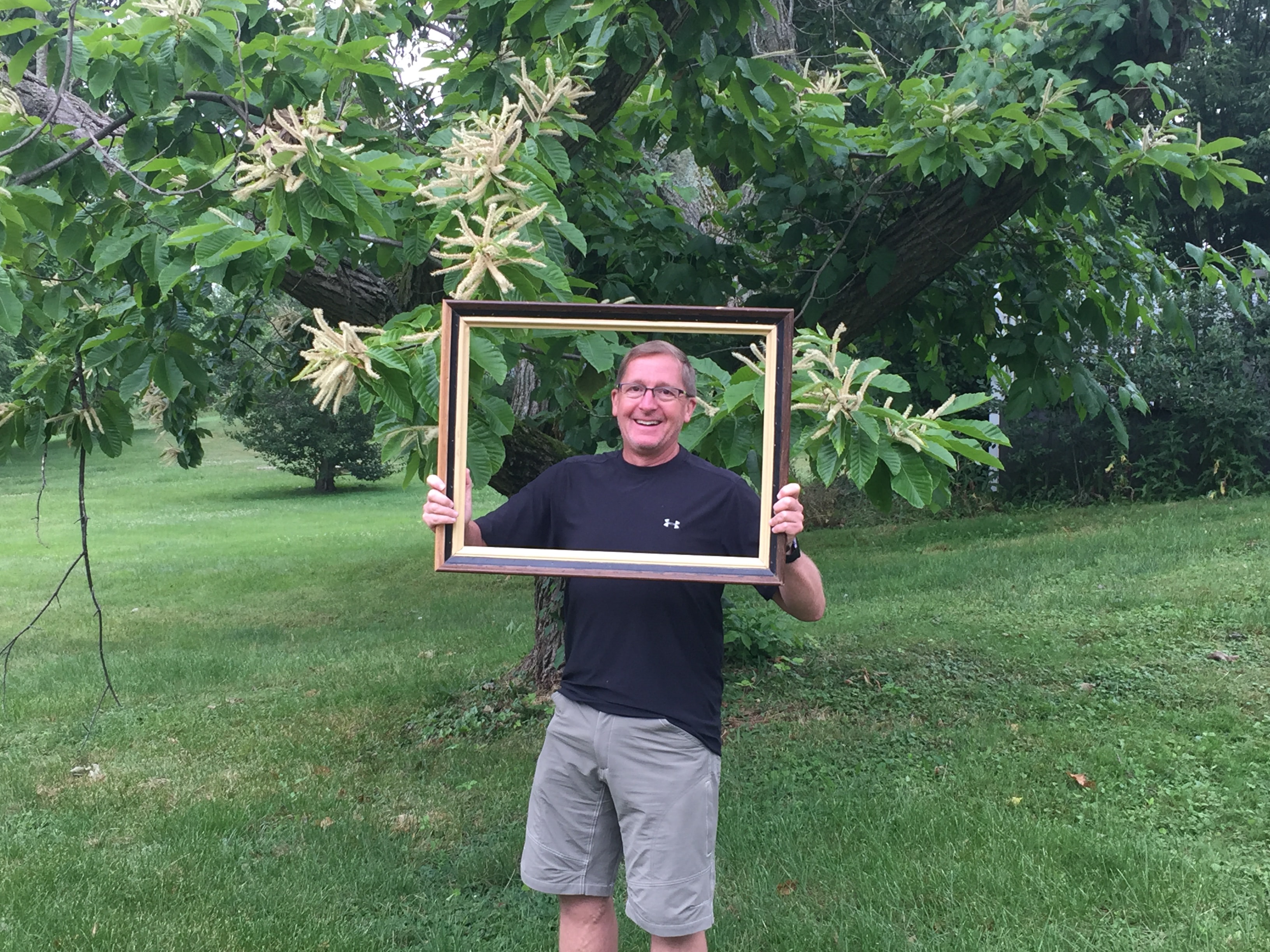 Smiling man in a black Under Armour shirt holding a large gold picture frame outdoors in a lush green garden beneath a flowering chestnut tree