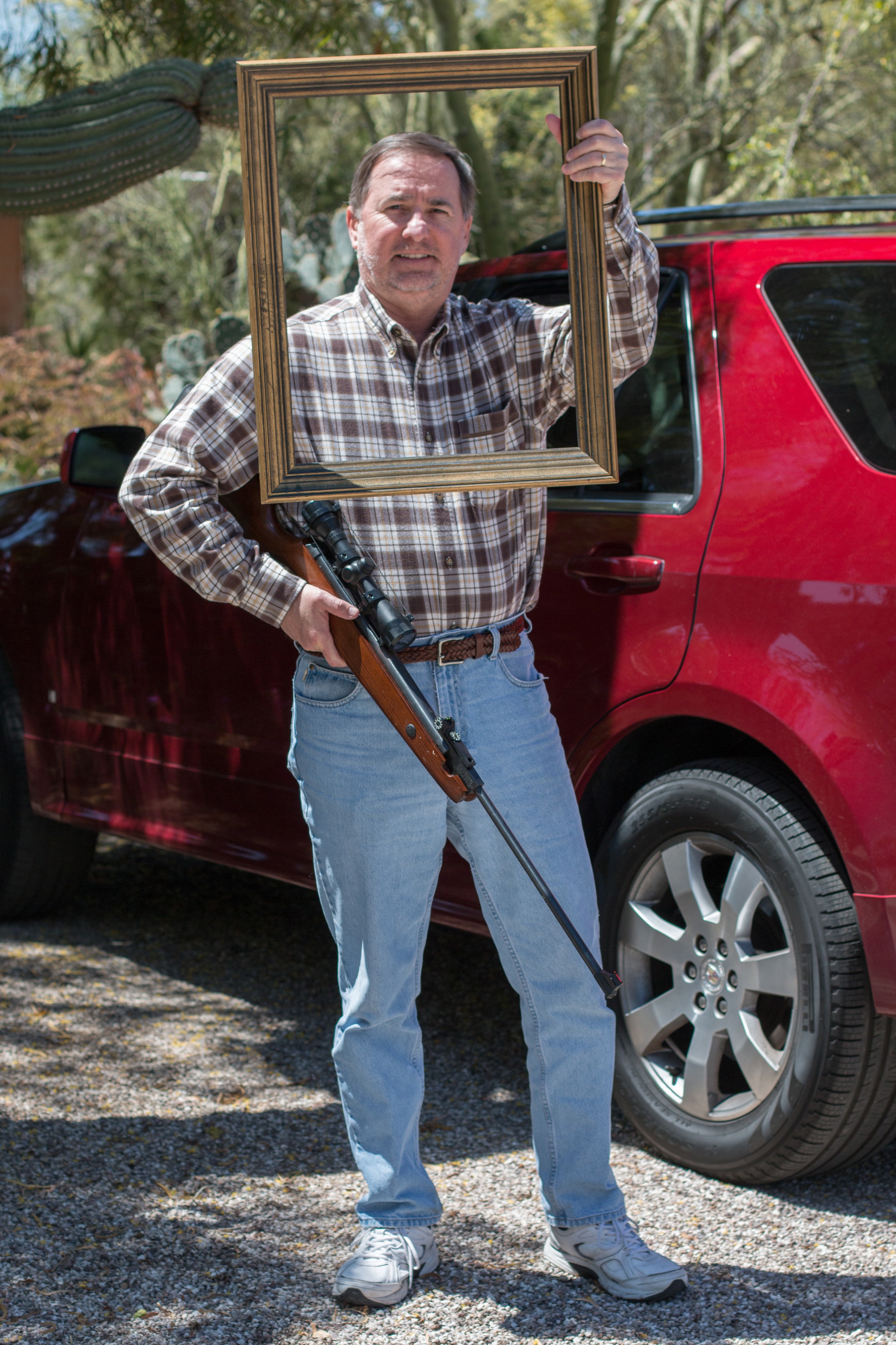 Man in a plaid shirt holding a gold picture frame with one hand and a scoped rifle with the other, standing in front of a red SUV with saguaro cactus behind him