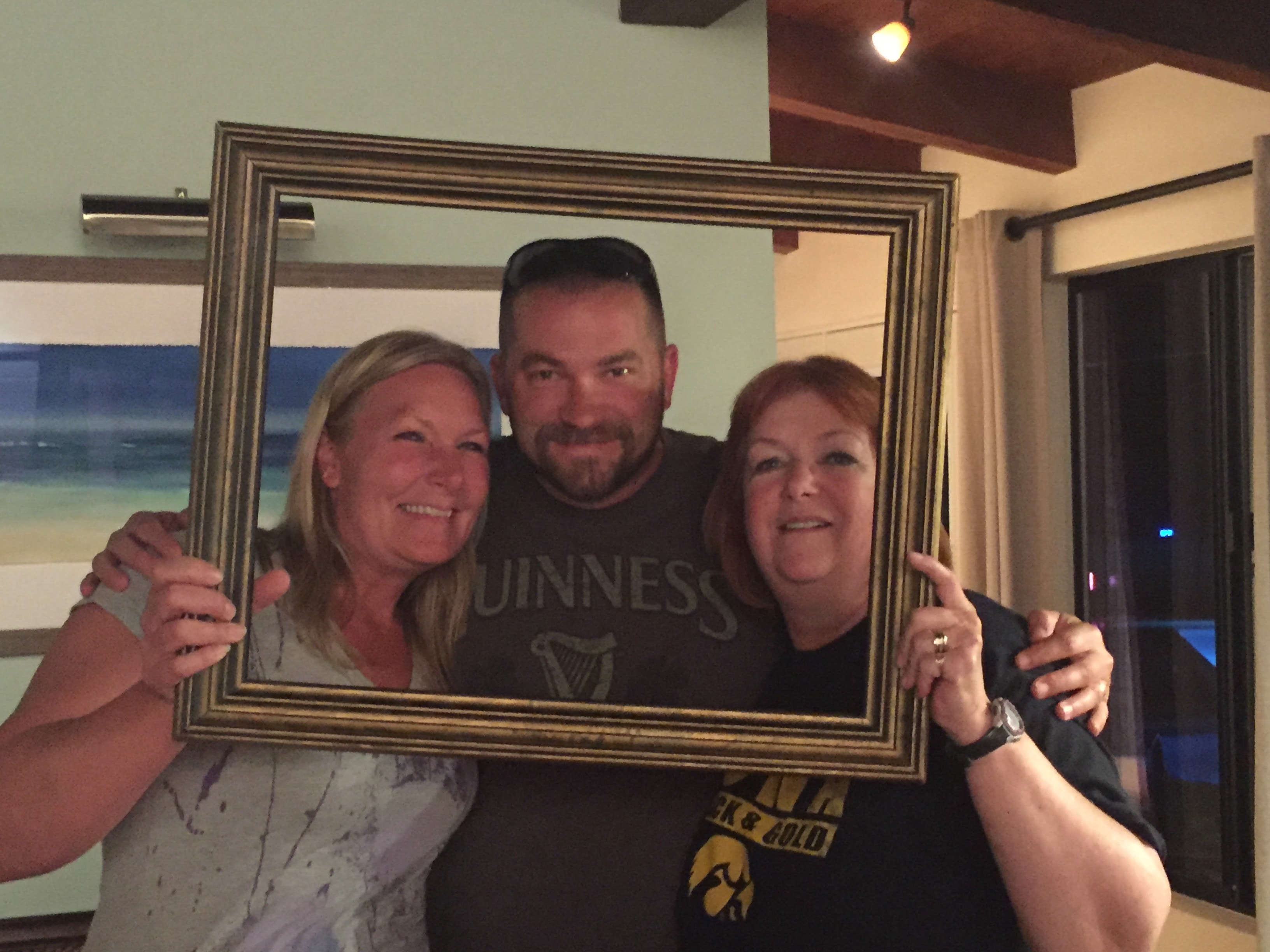 Three people — two women and a man in a Guinness shirt — crowding together through a gold picture frame indoors with a beach painting on the wall behind them