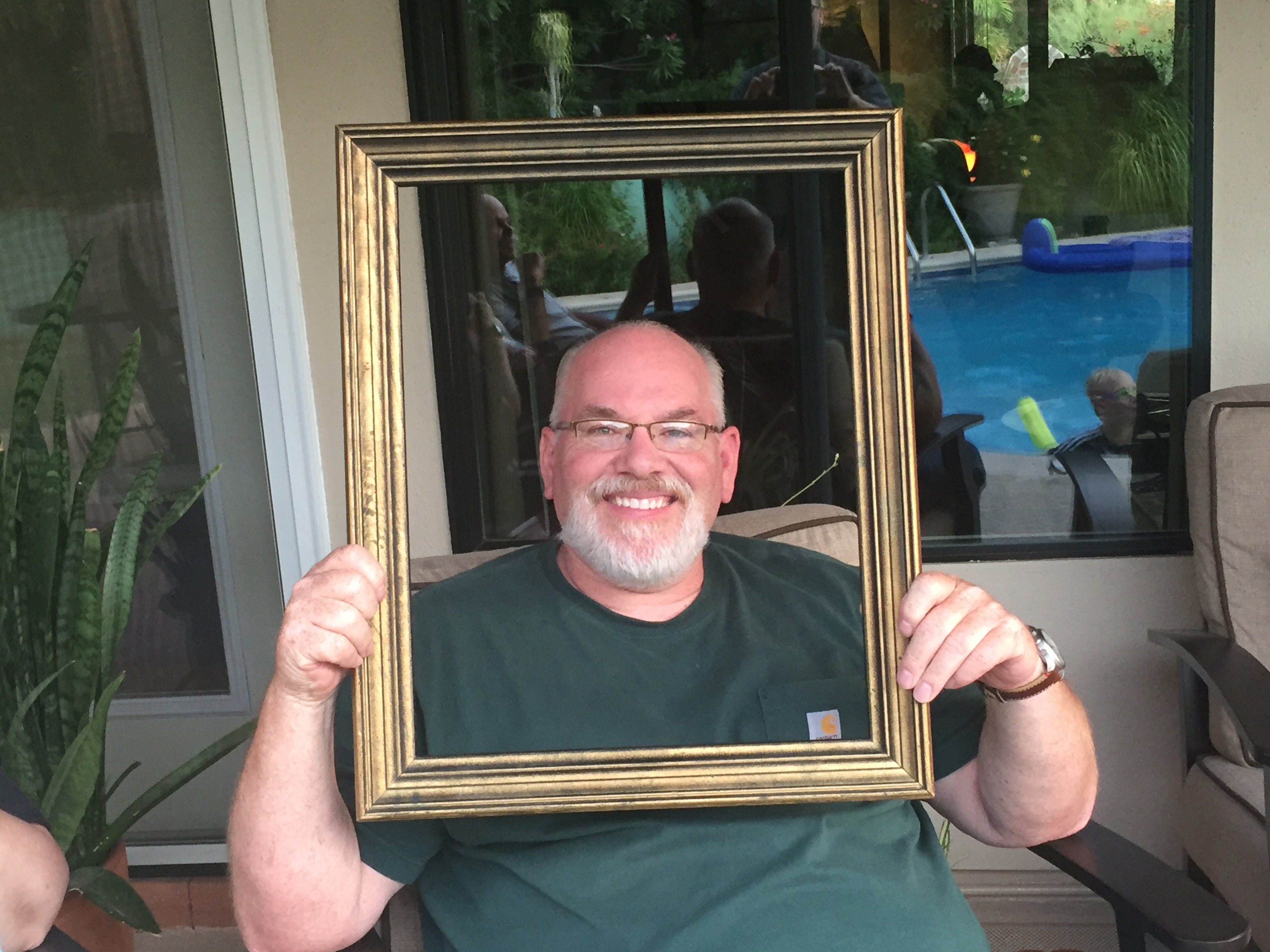 Bearded man in a green Carhartt shirt grinning broadly through a gold picture frame on a covered patio with a pool and party guests reflected in the glass behind him