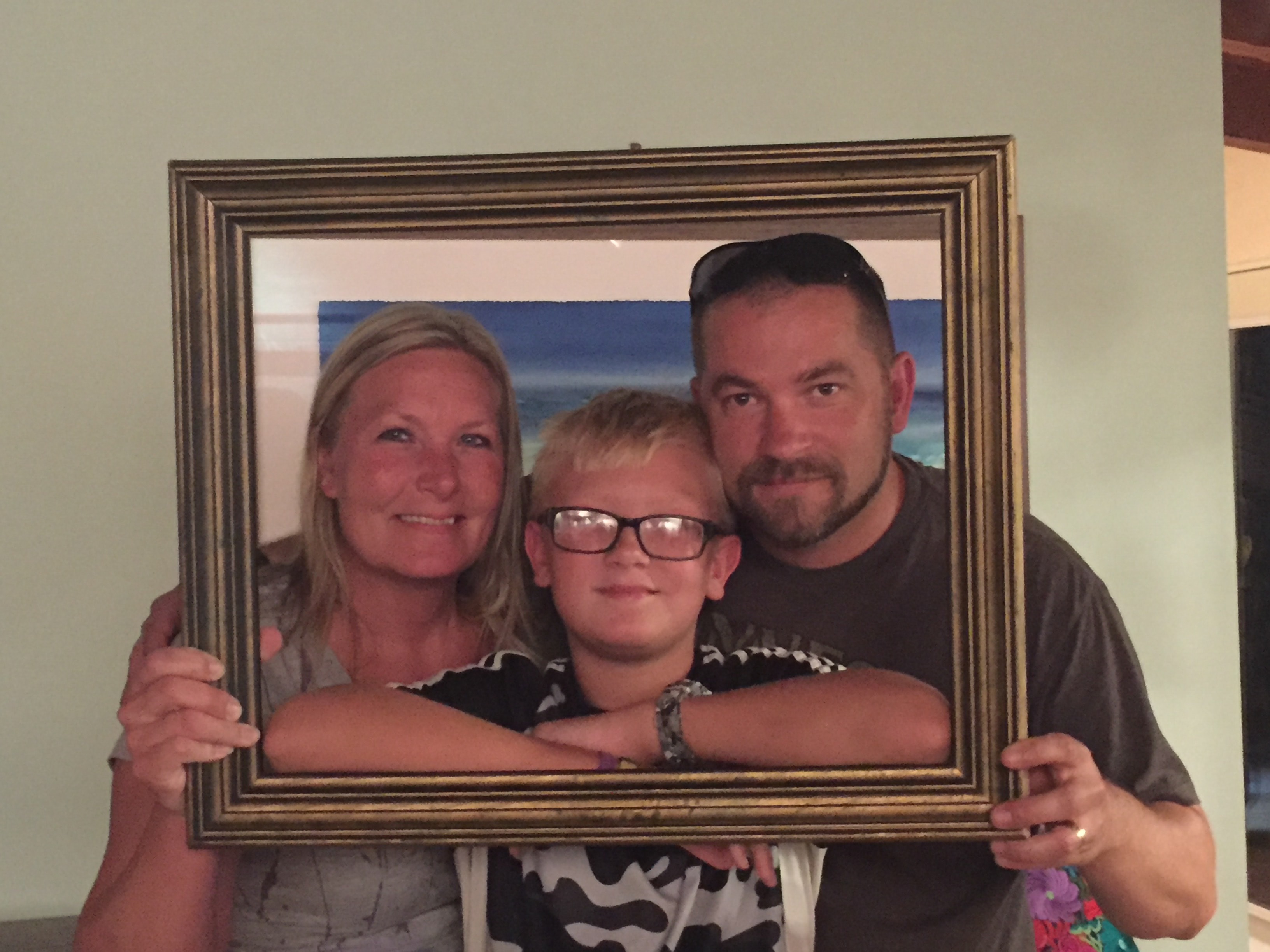 Mom, dad, and young boy with glasses smiling together through a gold picture frame indoors with a beach painting on the wall behind them