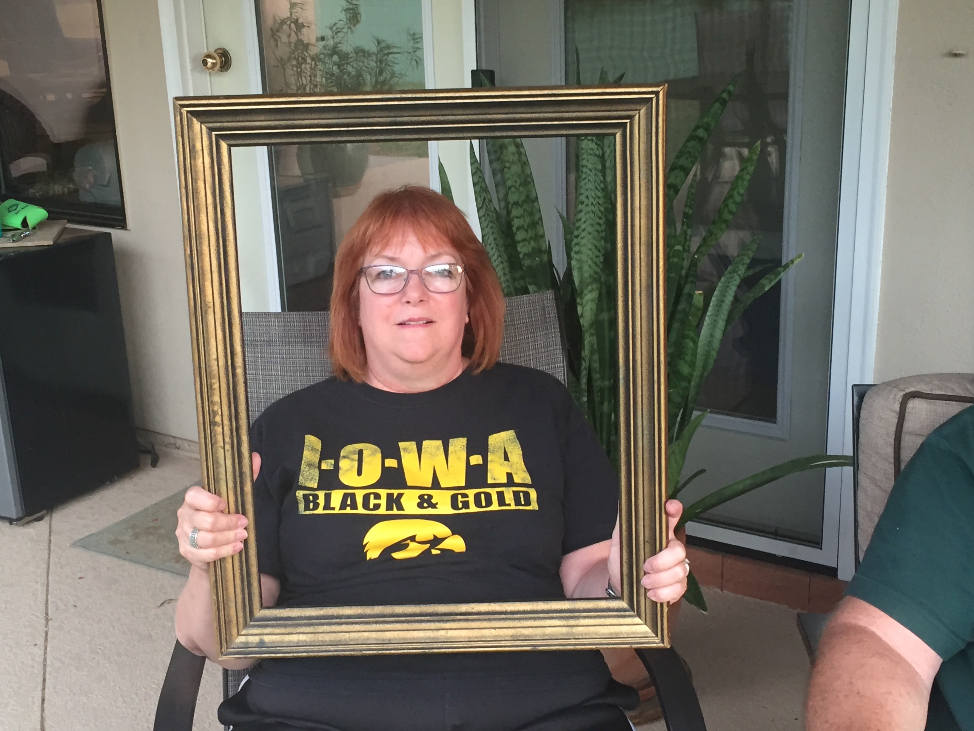 Woman in an Iowa Hawkeyes Black and Gold shirt sitting on a patio chair and holding a gold picture frame with snake plants behind her
