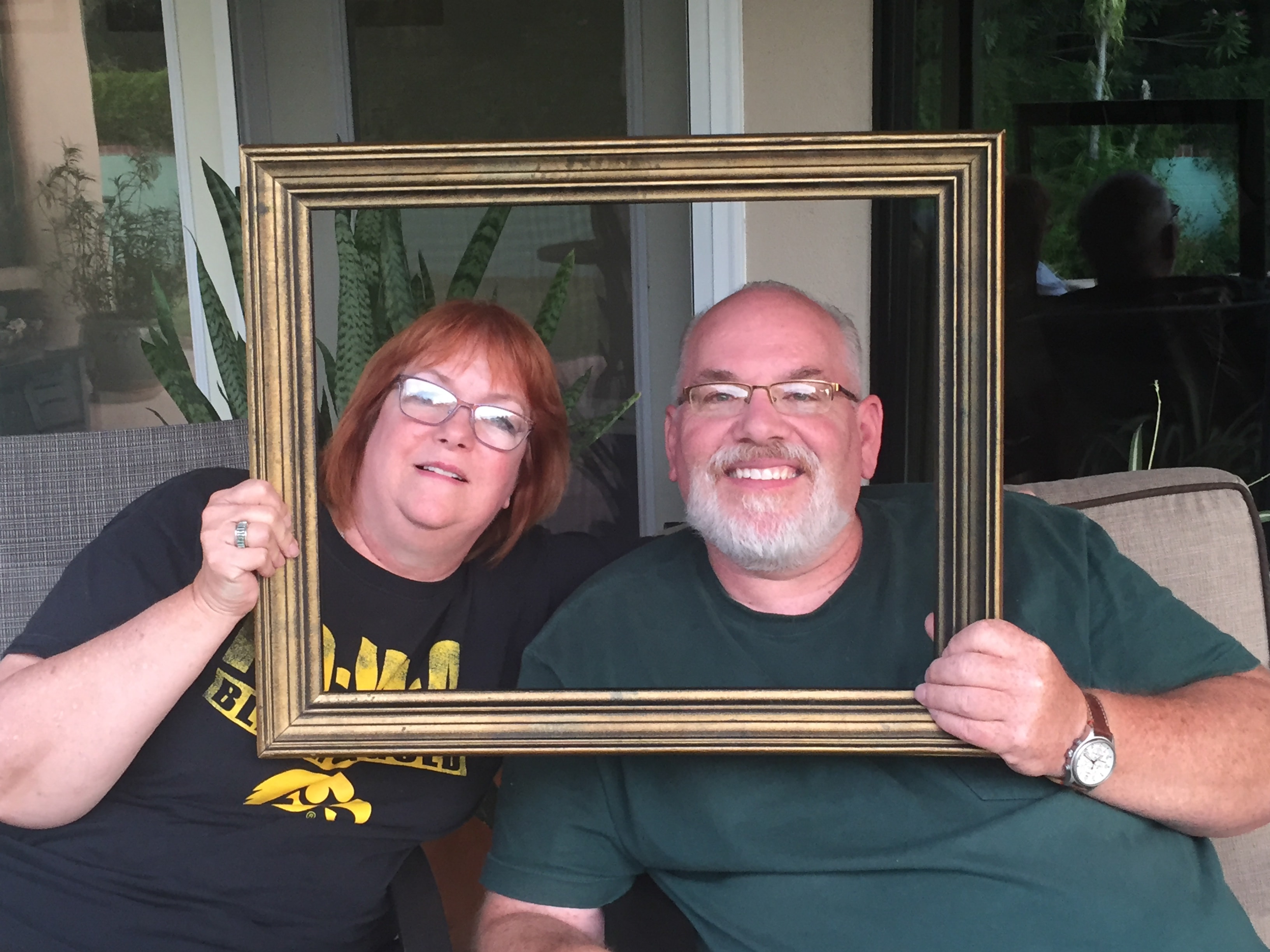 Woman in an Iowa Hawkeyes shirt and bearded man in a green Carhartt shirt sitting side by side and holding a gold picture frame together on a covered patio