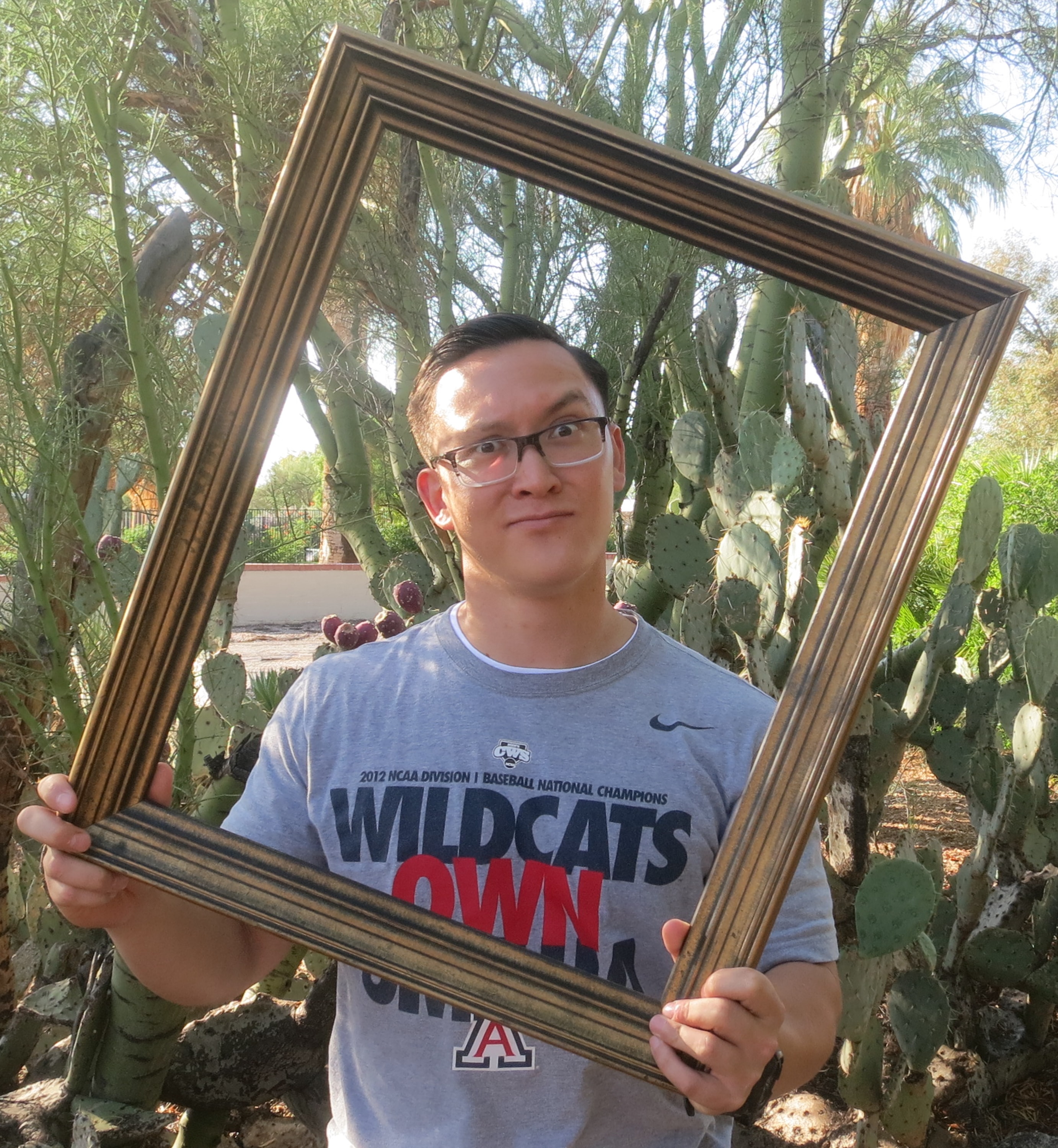 Young man in glasses and a Wildcats Own 2012 NCAA Baseball Champions shirt holding a large gold picture frame outdoors in front of prickly pear and saguaro cactus
