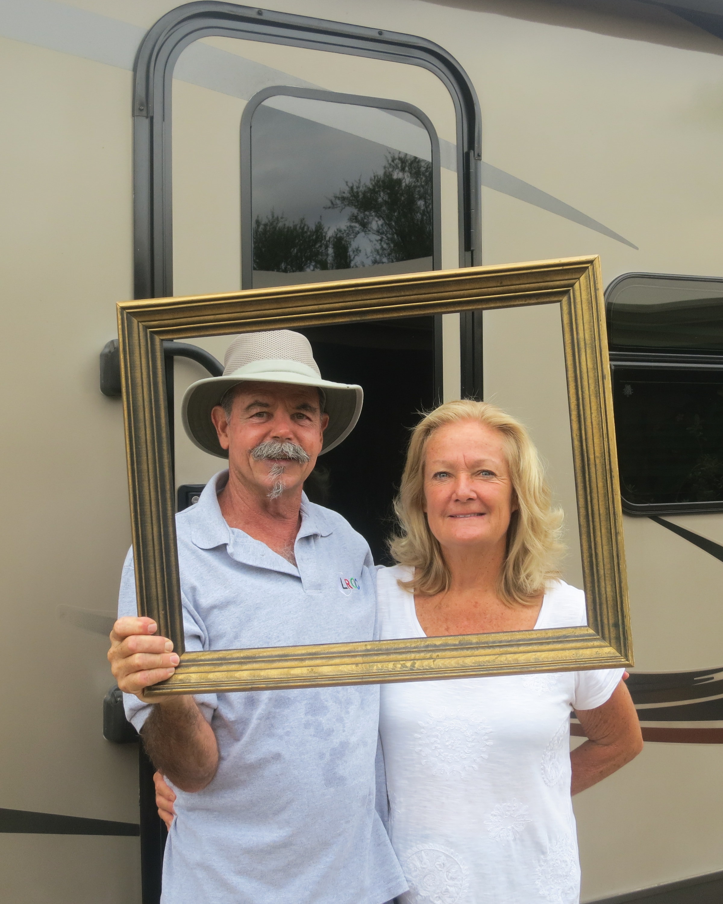 Man in a wide-brim hat and woman with blonde hair smiling together through a gold picture frame in front of a large RV